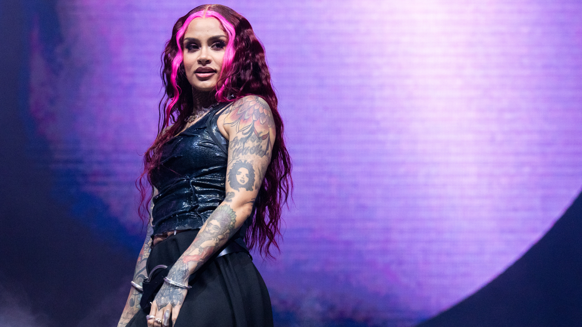Kehlani performing on stage, wearing a black outfit. Her long hair is styled with pink highlights, and she has visible tattoos.