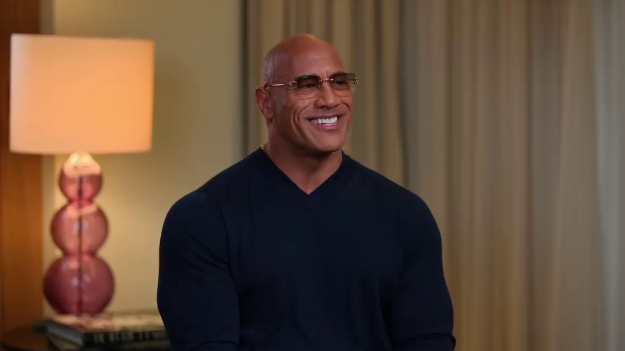 Dwayne "The Rock" Johnson smiling, wearing glasses and a dark shirt, sitting in a room with a lamp in the background.