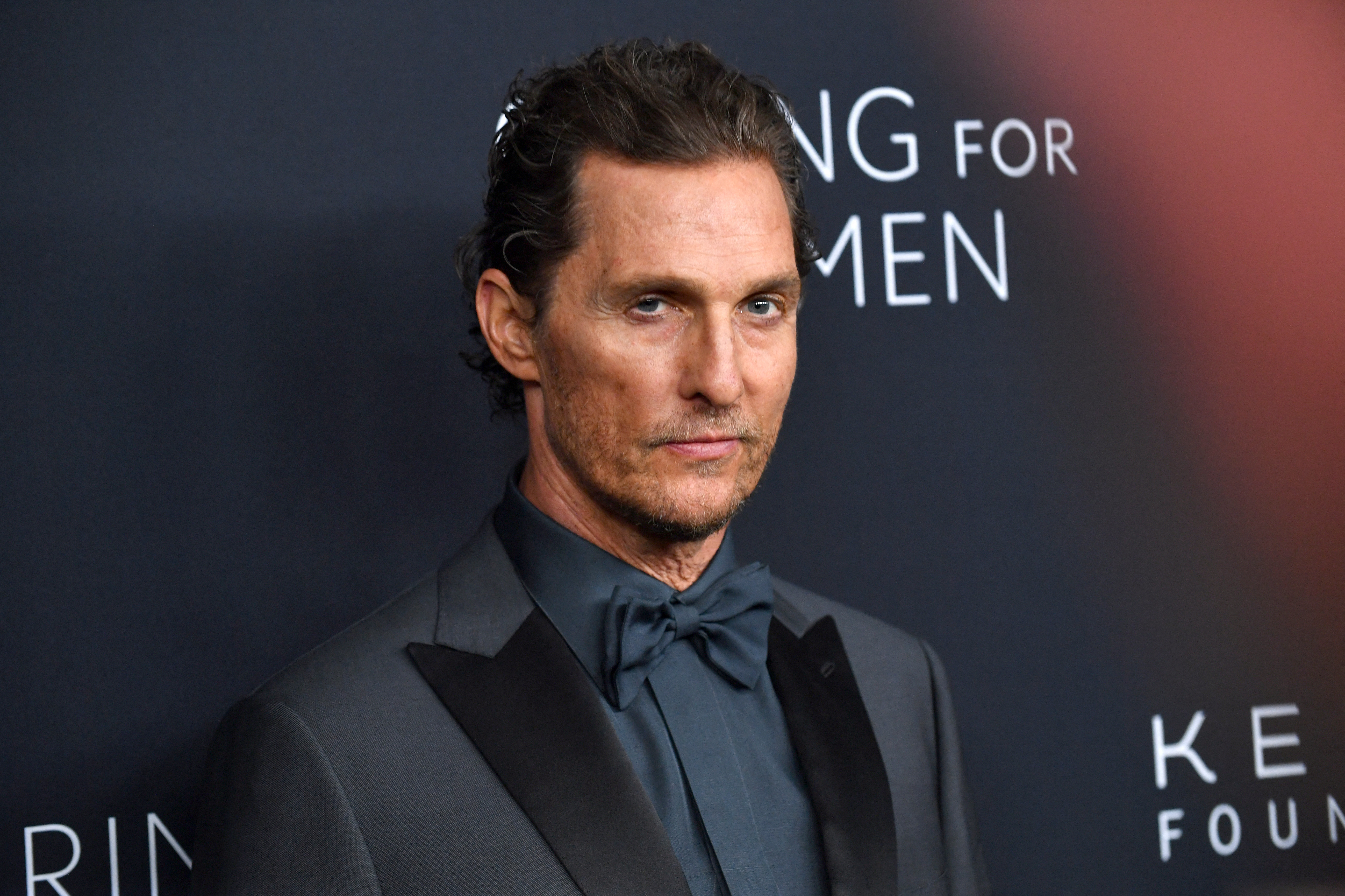 Matthew McConaughey in a dark suit and bow tie at an event, posing against a dark backdrop with partial text visible.