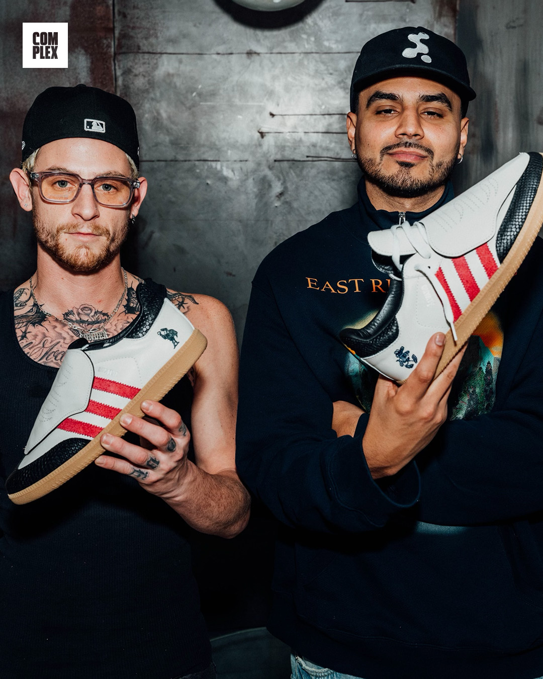 Two men holding sneakers with red stripes, one wearing glasses and a cap, the other in a black cap and sweatshirt.