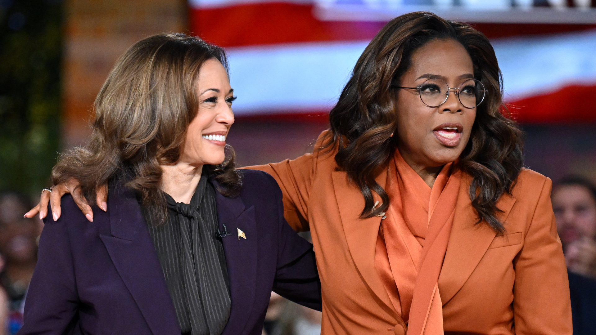 Kamala Harris and Oprah Winfrey stand together, smiling and talking. Kamala wears a dark suit, and Oprah wears an orange outfit.