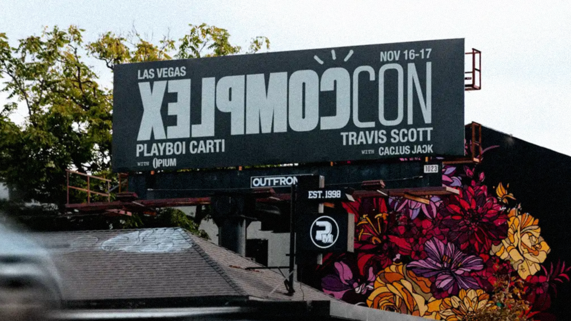 Billboard for ComplexCon in Las Vegas featuring Playboi Carti and Travis Scott, with colorful floral mural in the background.