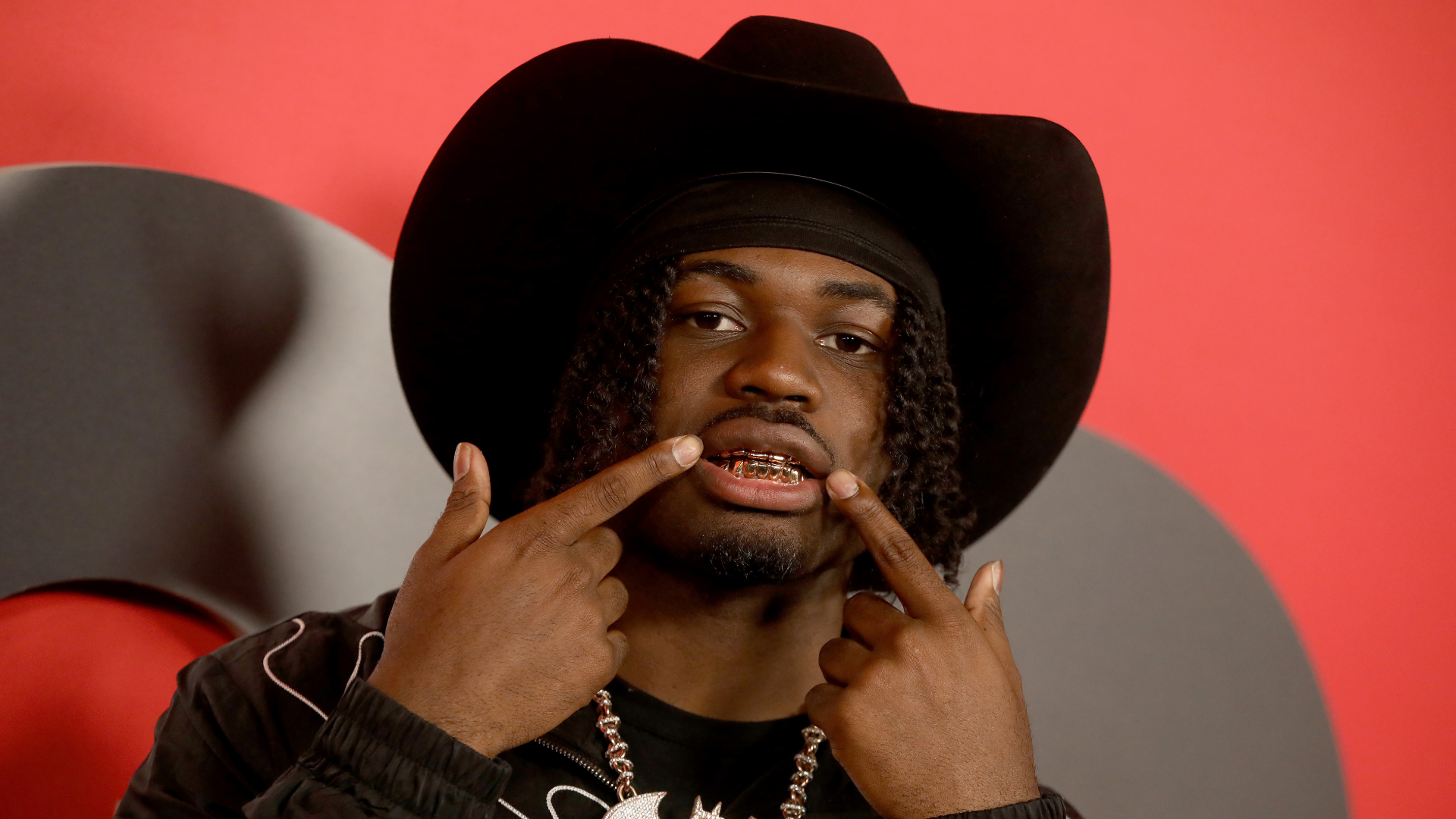 4Batz wearing a black cowboy hat shows off his gold grillz, pointing at them with both hands against a red background.