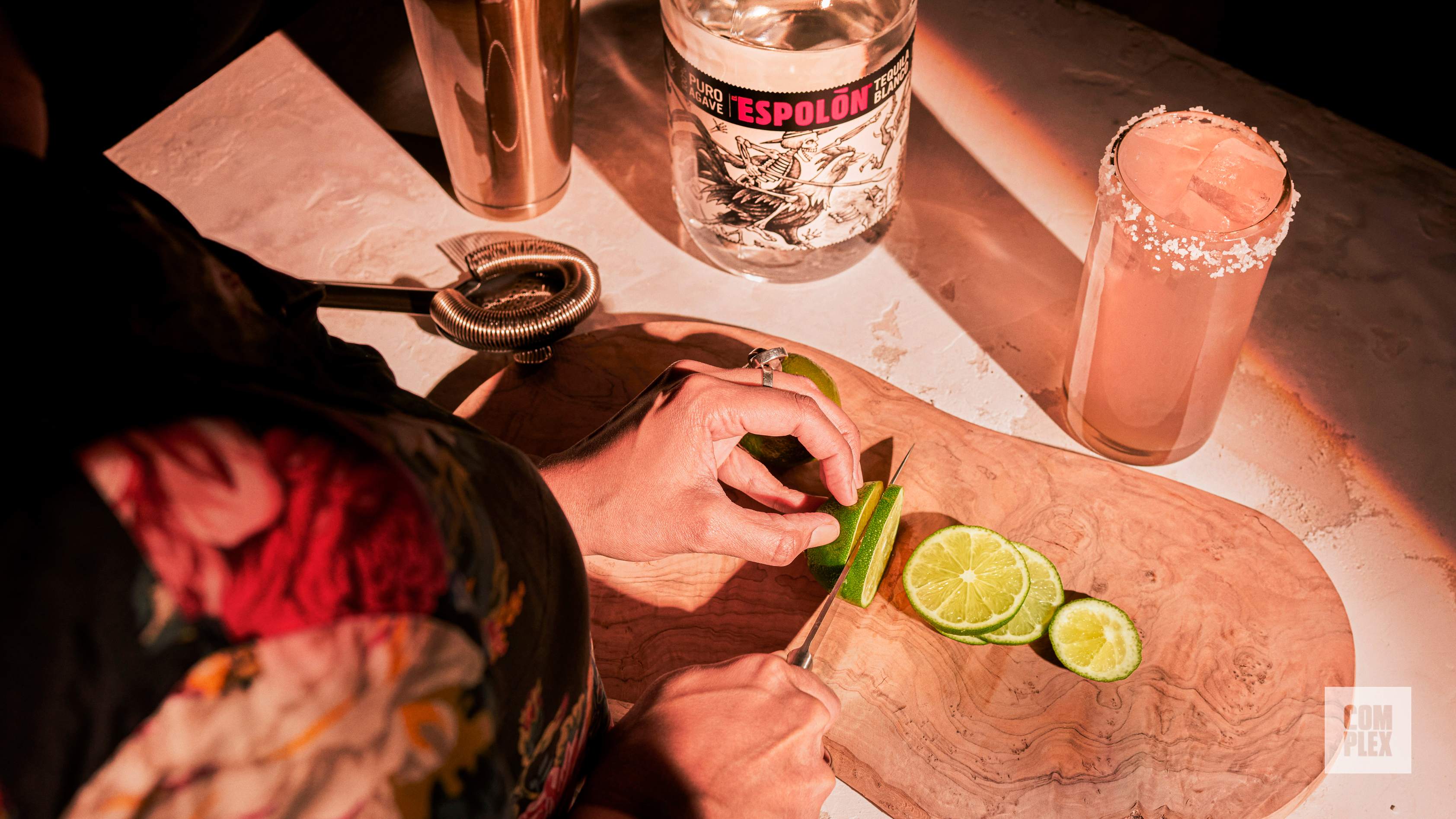 A person slices limes on a wooden board. Nearby are a bottle of Espolón tequila, a cocktail shaker, and a salt-rimmed drink.