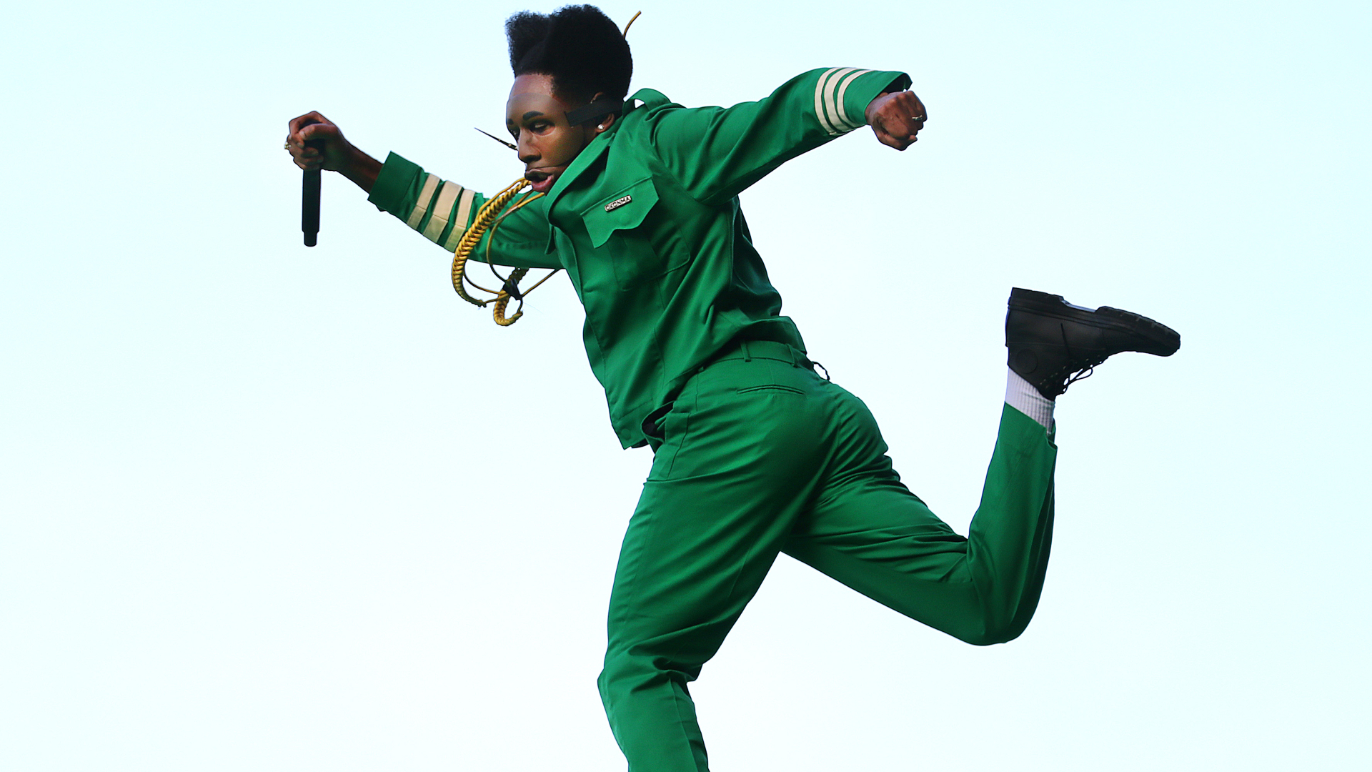 Tyler in a green suit mid-jump, holding a microphone, with an energetic pose against a plain background.