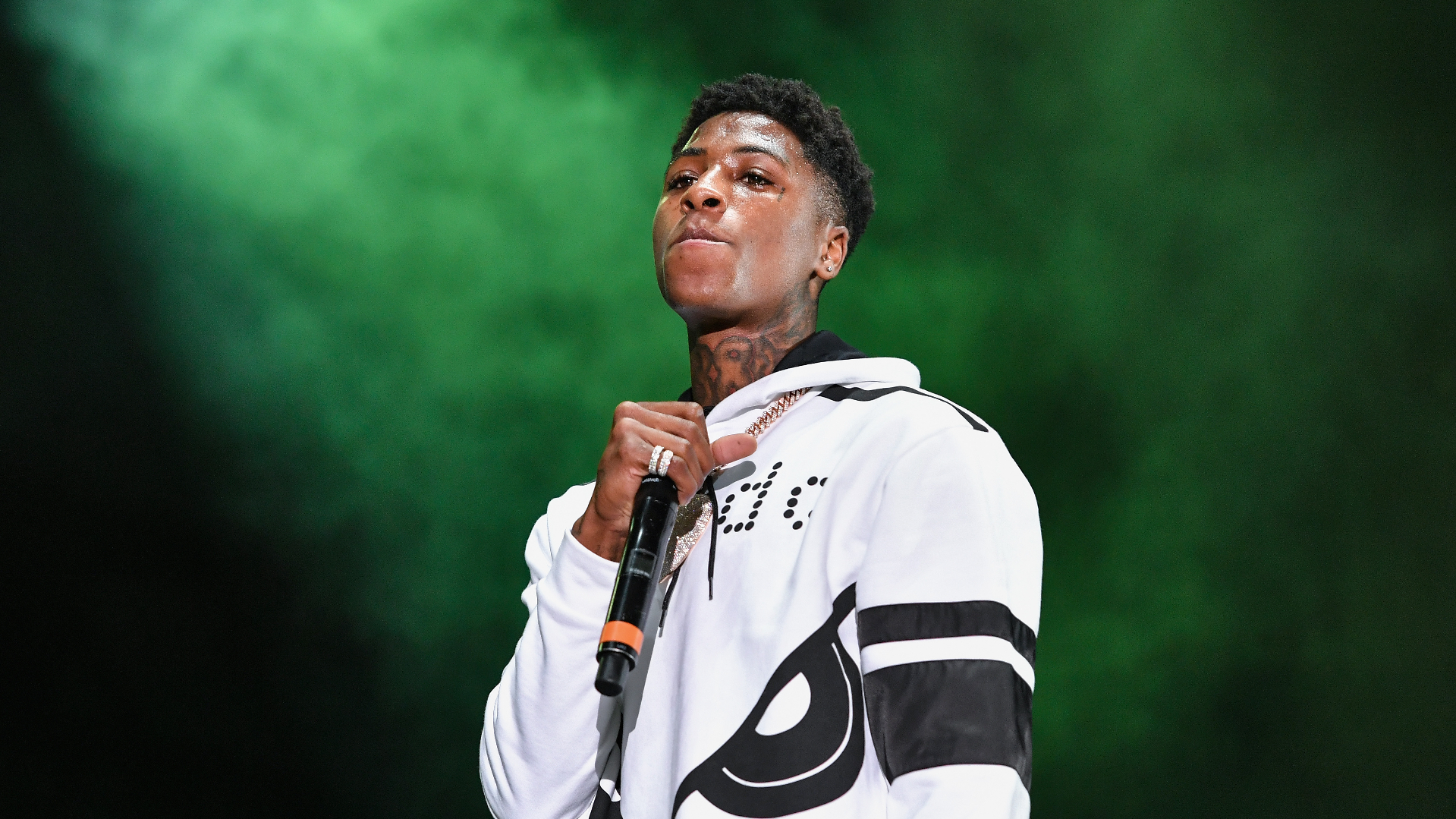 A man in a white hoodie holding a microphone on stage, with a green background.