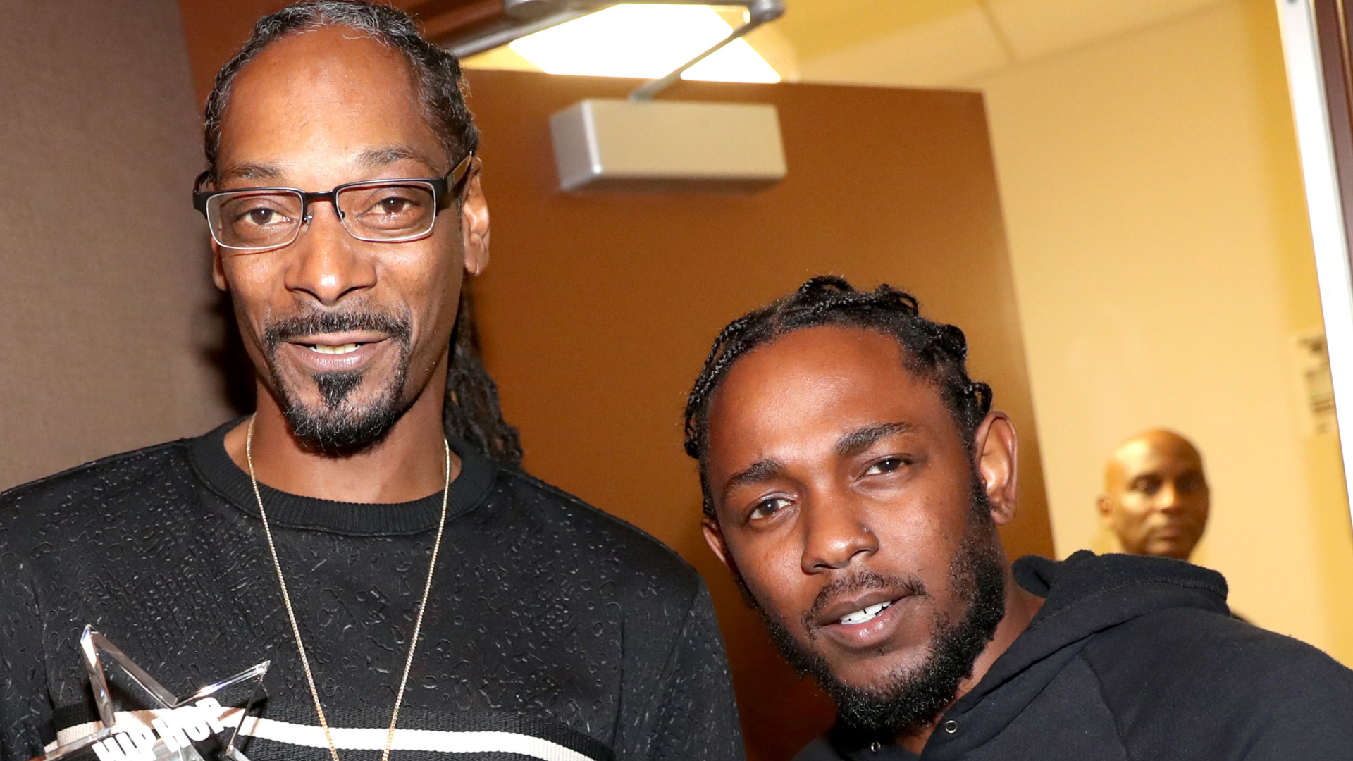 Snoop Dogg and Kendrick Lamar smiling together indoors, with another person in the background.