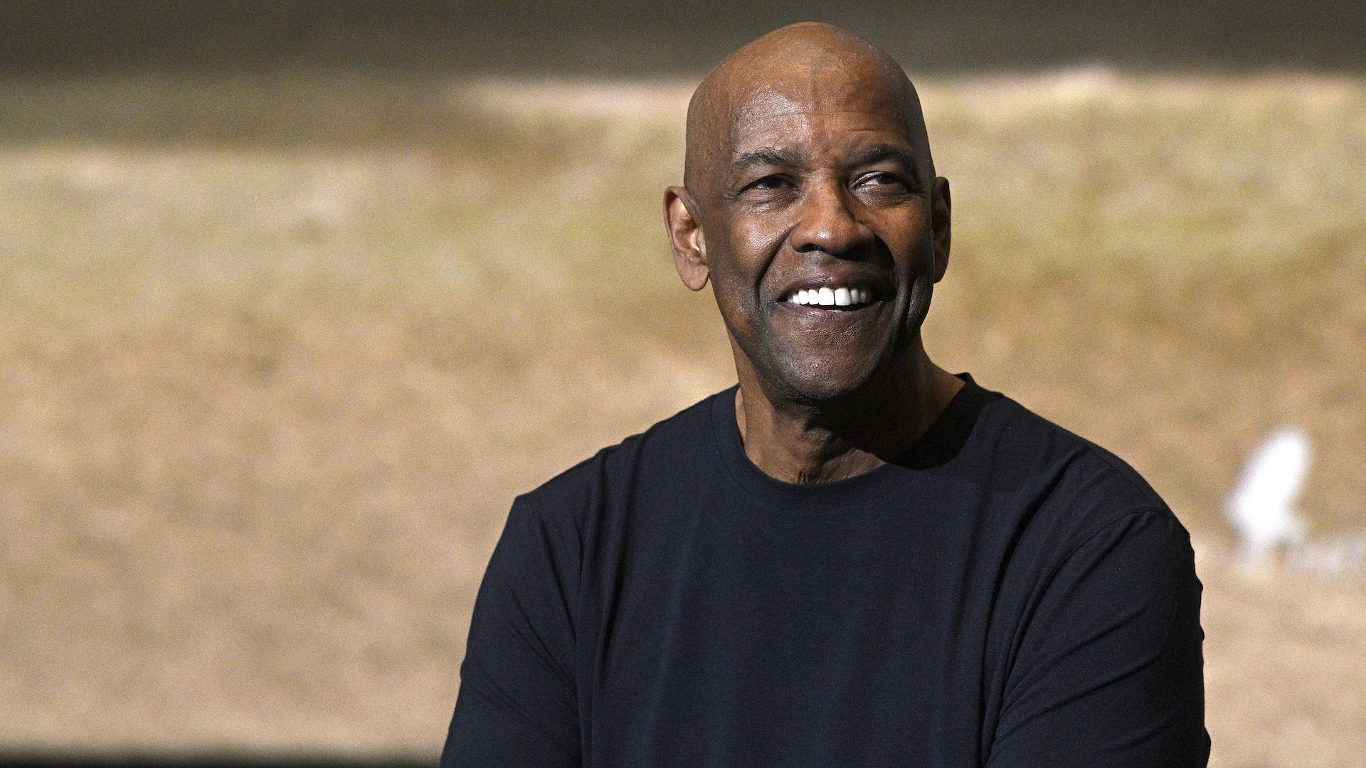 Denzel Washington smiling, wearing a black shirt, against a blurred background.