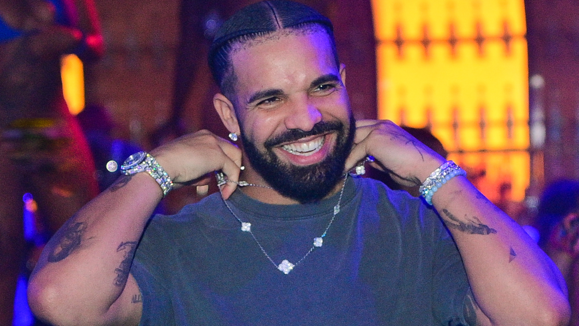 Drake smiling, wearing a black shirt, diamond necklace, and bracelets. Background is a dimly lit, colorful venue.