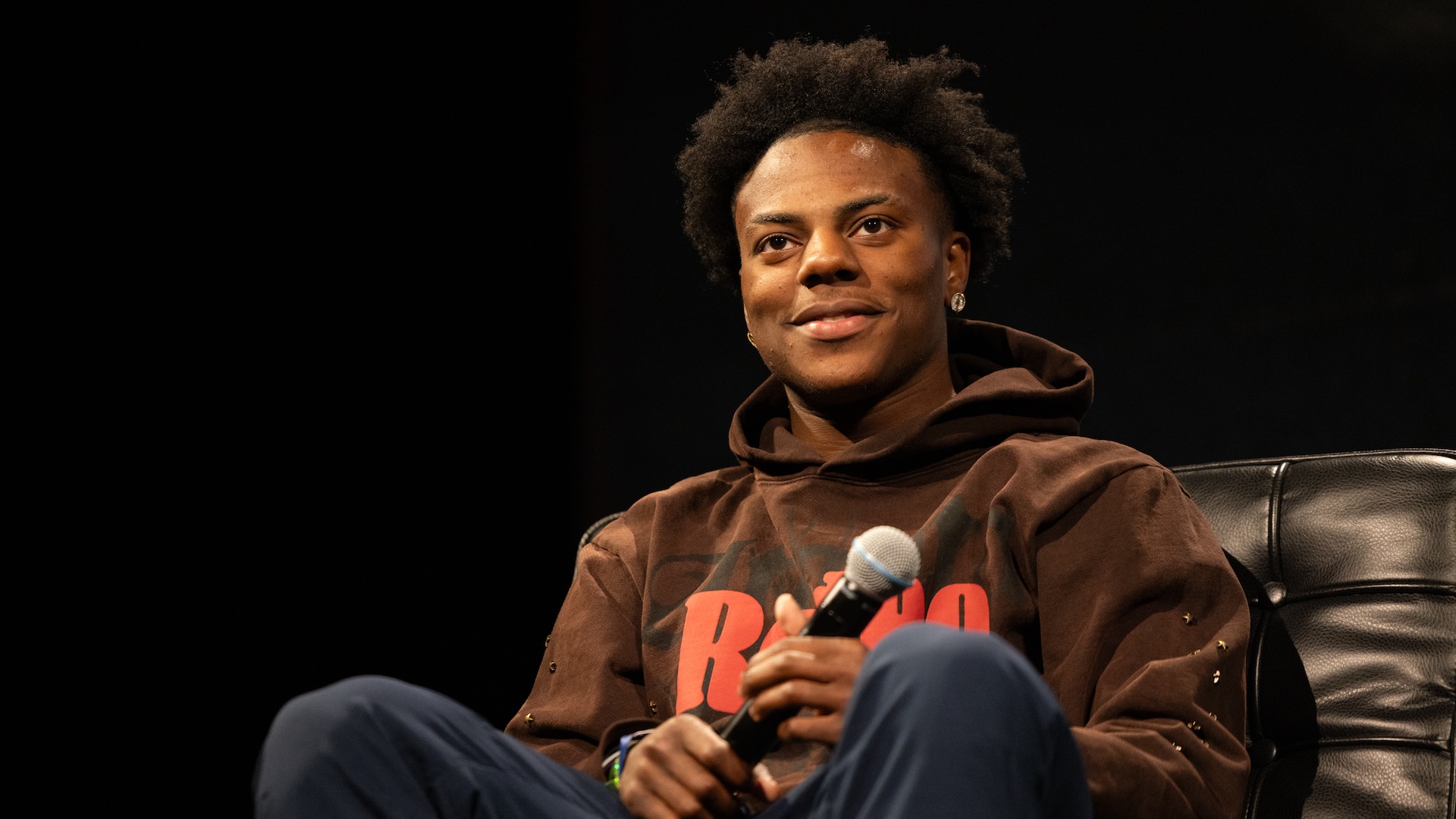 IShowSpeed with short curly hair, wearing a brown hoodie, sits on a chair holding a microphone.