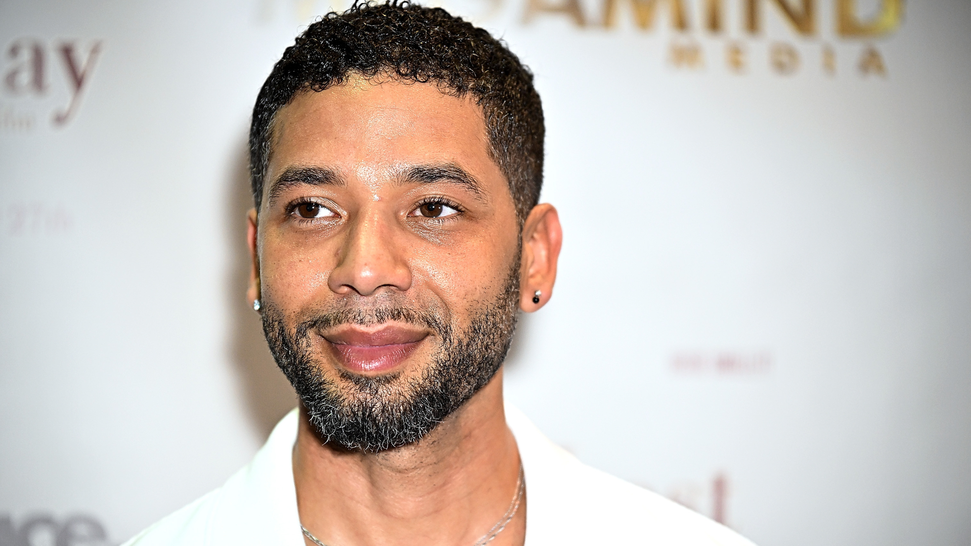 Jussie Smollett at a media event, wearing a white shirt, with a neutral background.