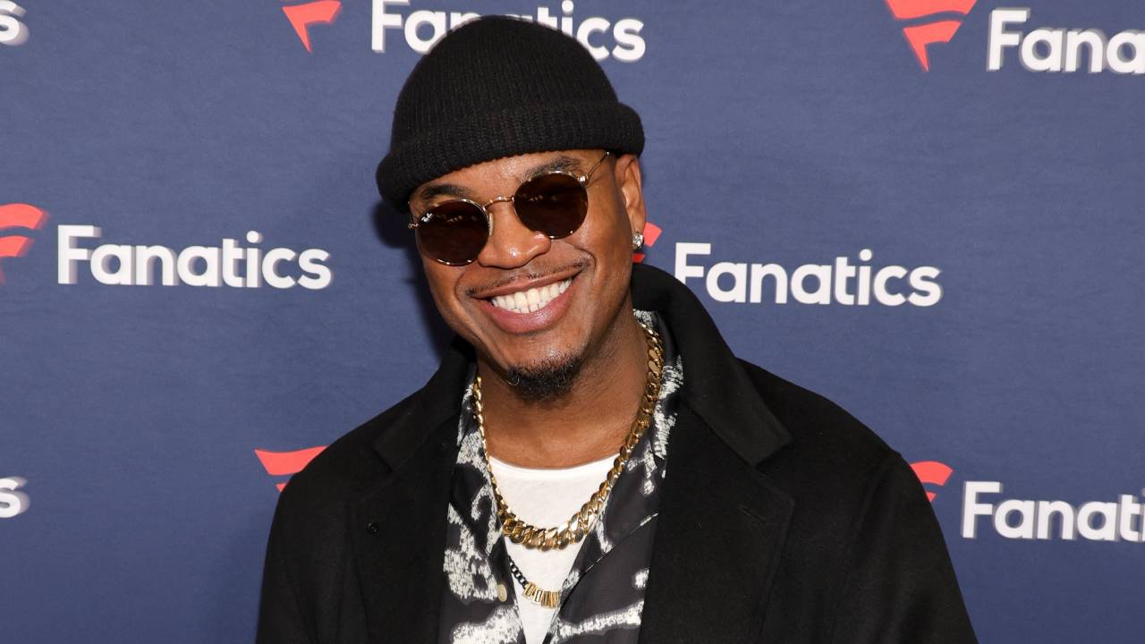Ne-Yo smiles at an event, wearing sunglasses, a black beanie, and a stylish outfit with gold chains.