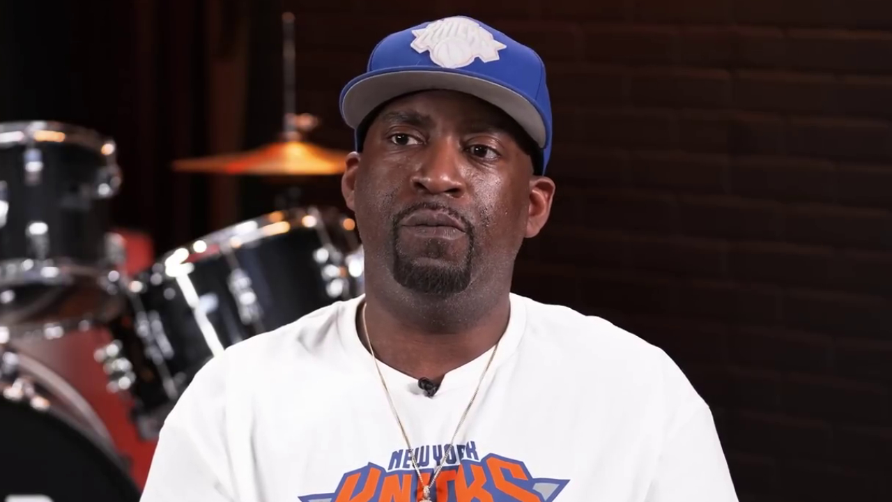 Tony Yayo wearing a New York Knicks shirt and a blue cap sits in front of a drum set.
