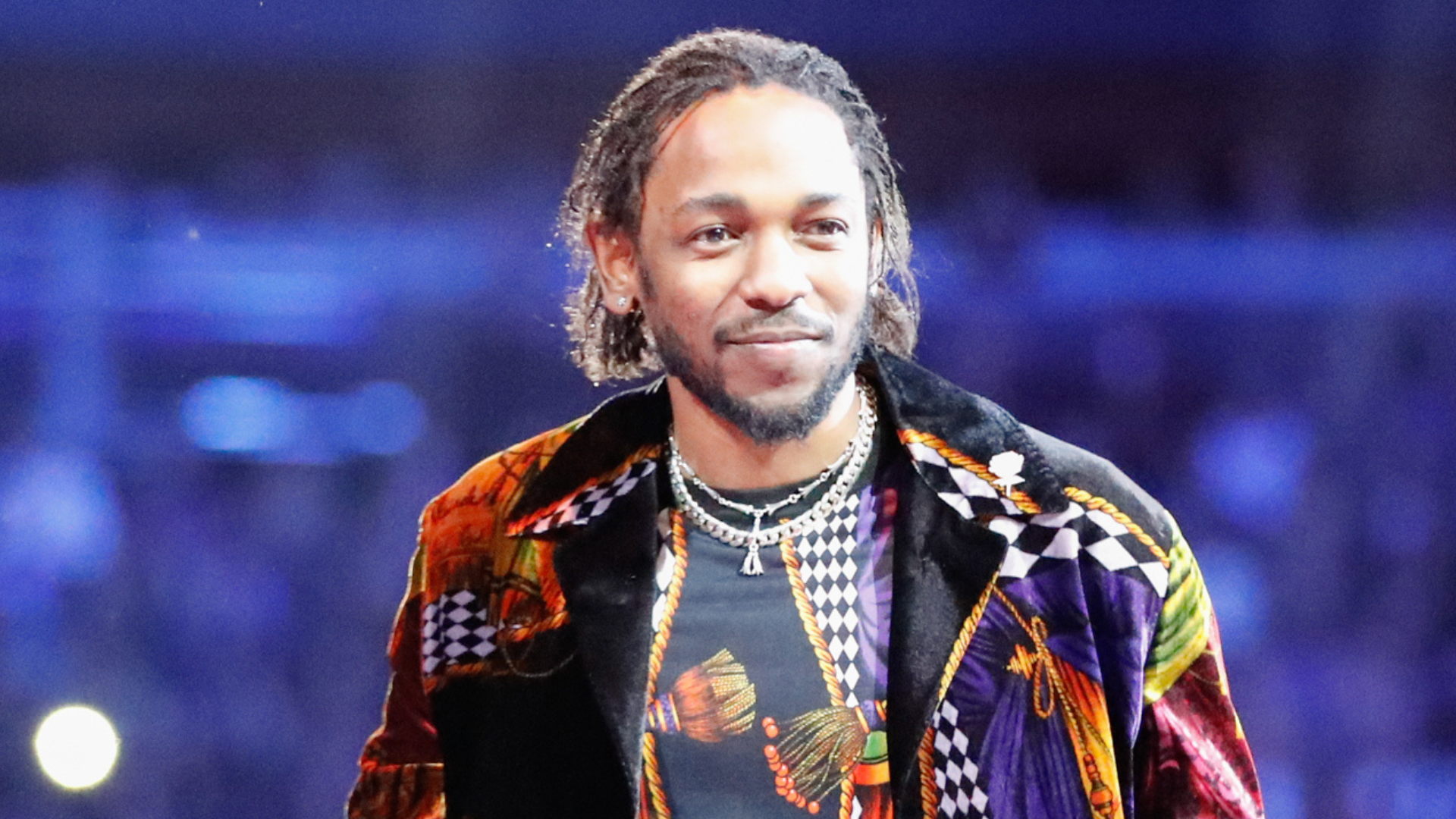 Kendrick Lamar wearing a colorful, patterned jacket on stage, smiling with braided hair and a necklace.