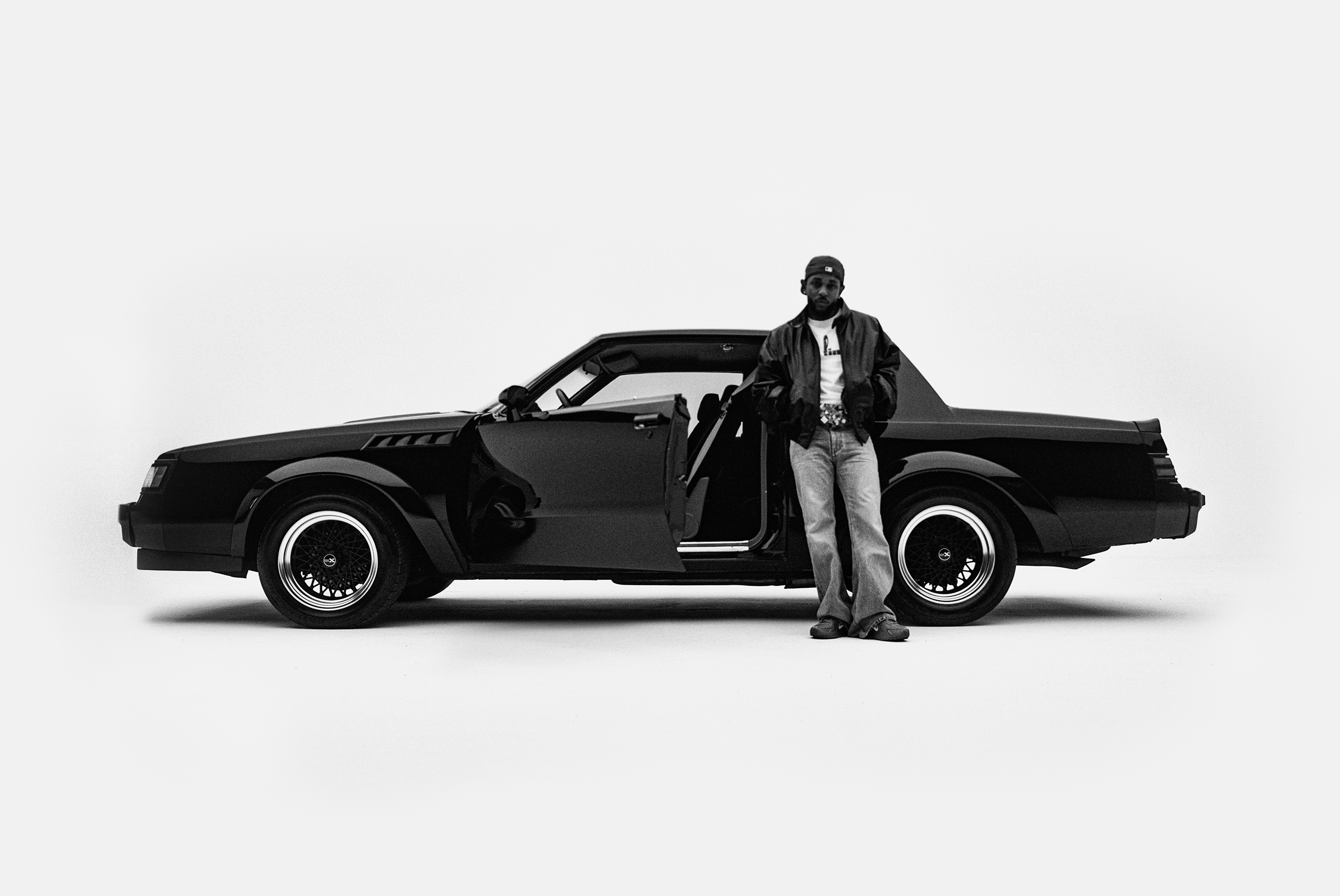 Kendrick Lamar stands beside a classic black car with open doors, wearing a cap and leather jacket, in a minimalist setting.