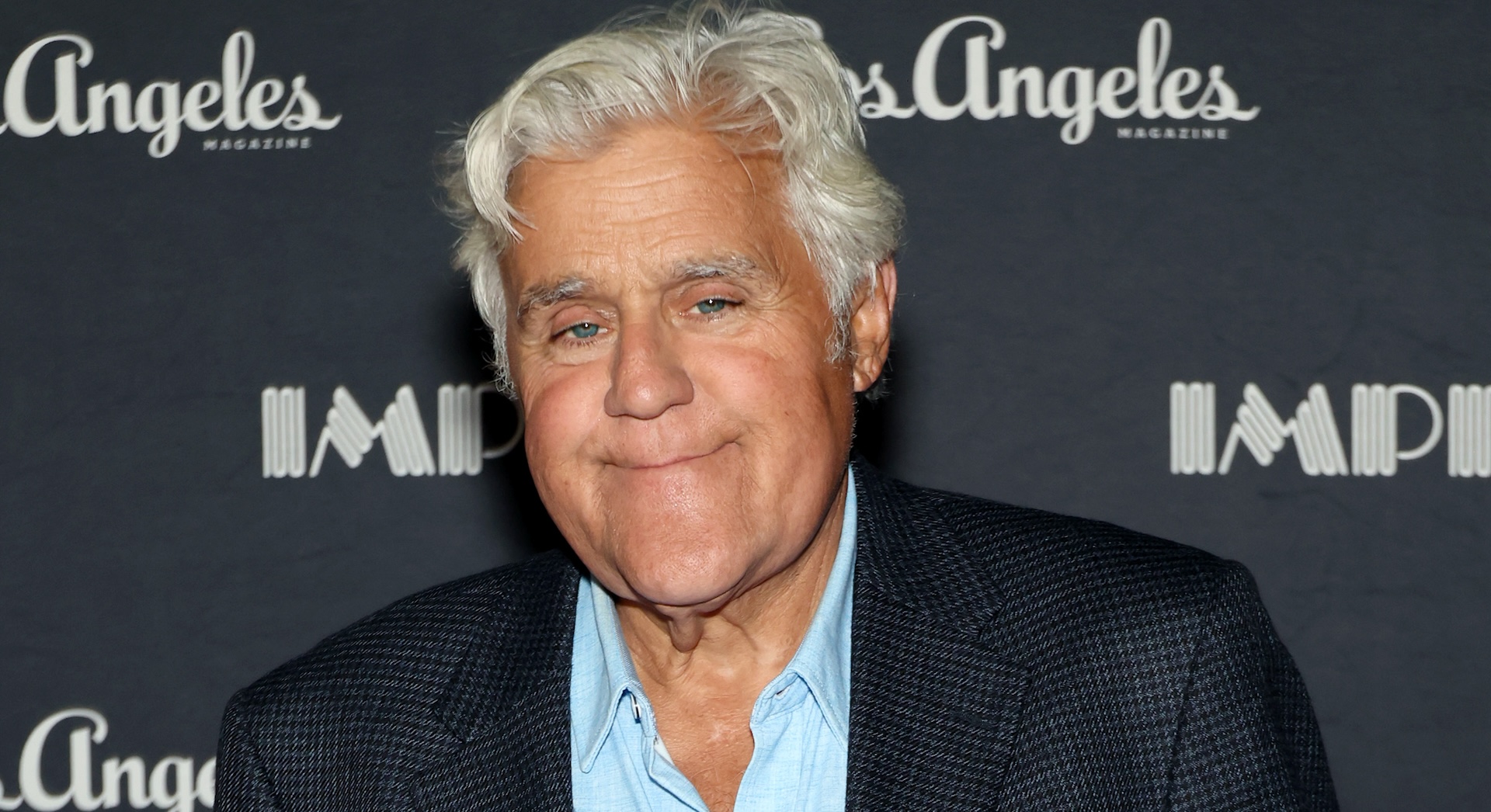 Jay Leno smiling in front of a backdrop with "Los Angeles Magazine" text. He's wearing a dark suit and light blue shirt.
