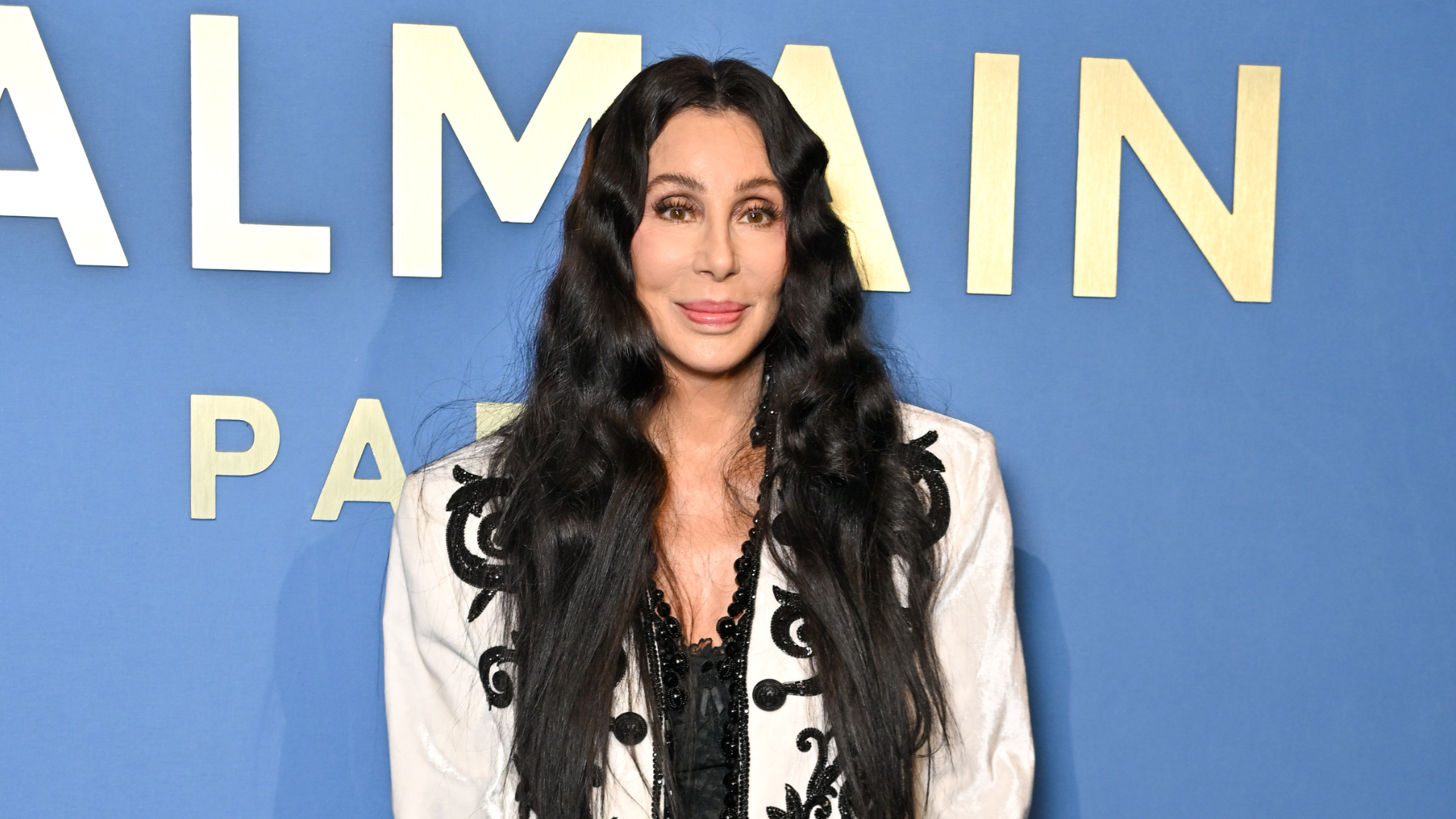 Cher in a black and white outfit with long wavy hair, standing in front of a blue background with "Balmain" written on it.
