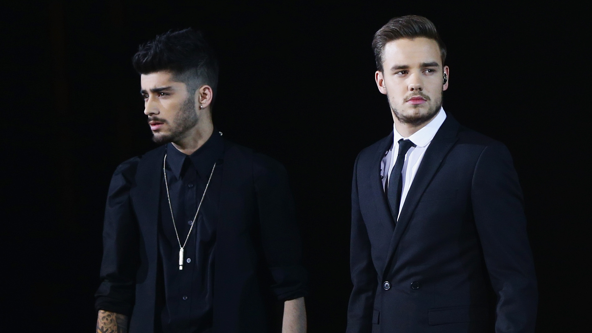 Zayn Malik and Liam Payne in formal attire, standing side by side against a dark background.