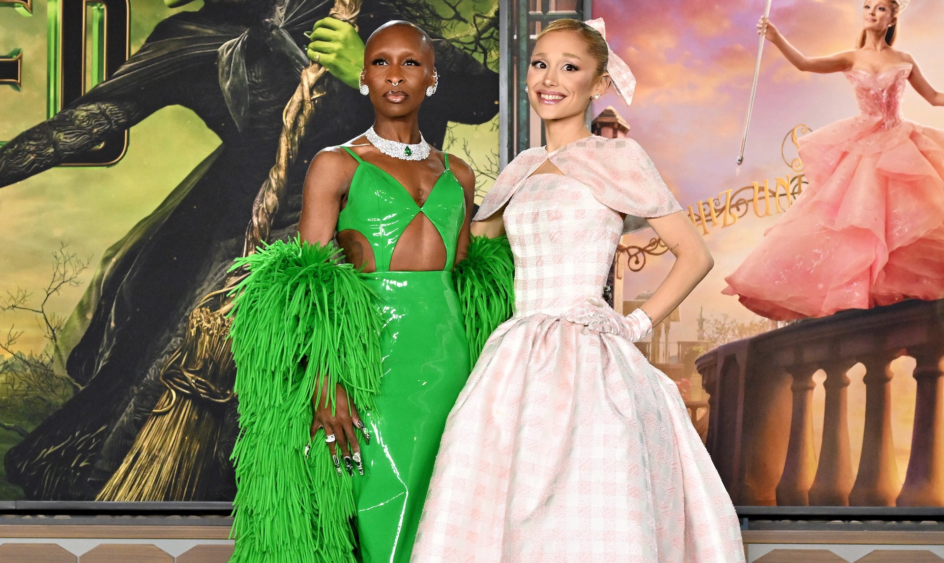 Cynthia Erivo in a green dress with feathers and Ariana Grande in a pink gown, posing together at an event.