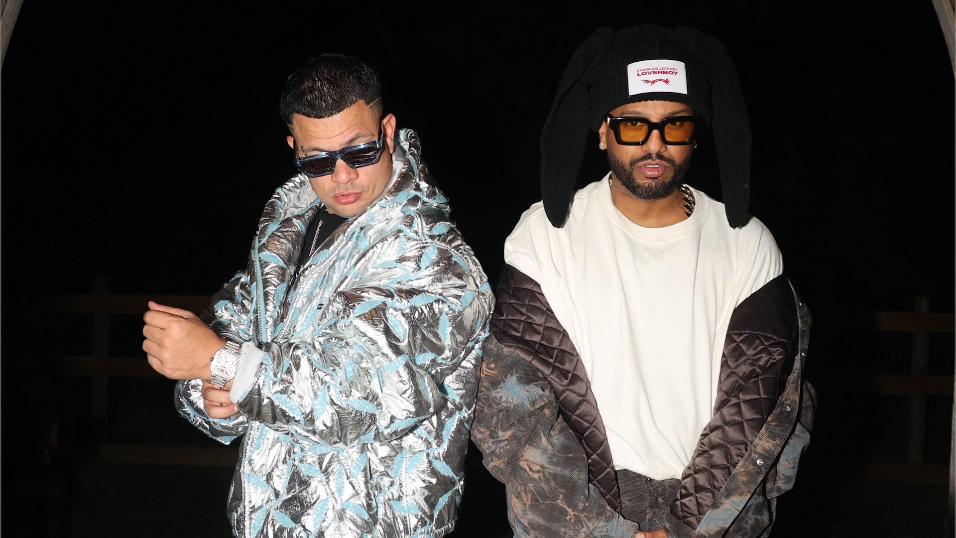 Jowell & Randy wearing stylish jackets and sunglasses, posing confidently. One has a metallic jacket, the other a quilted coat with a unique hat.