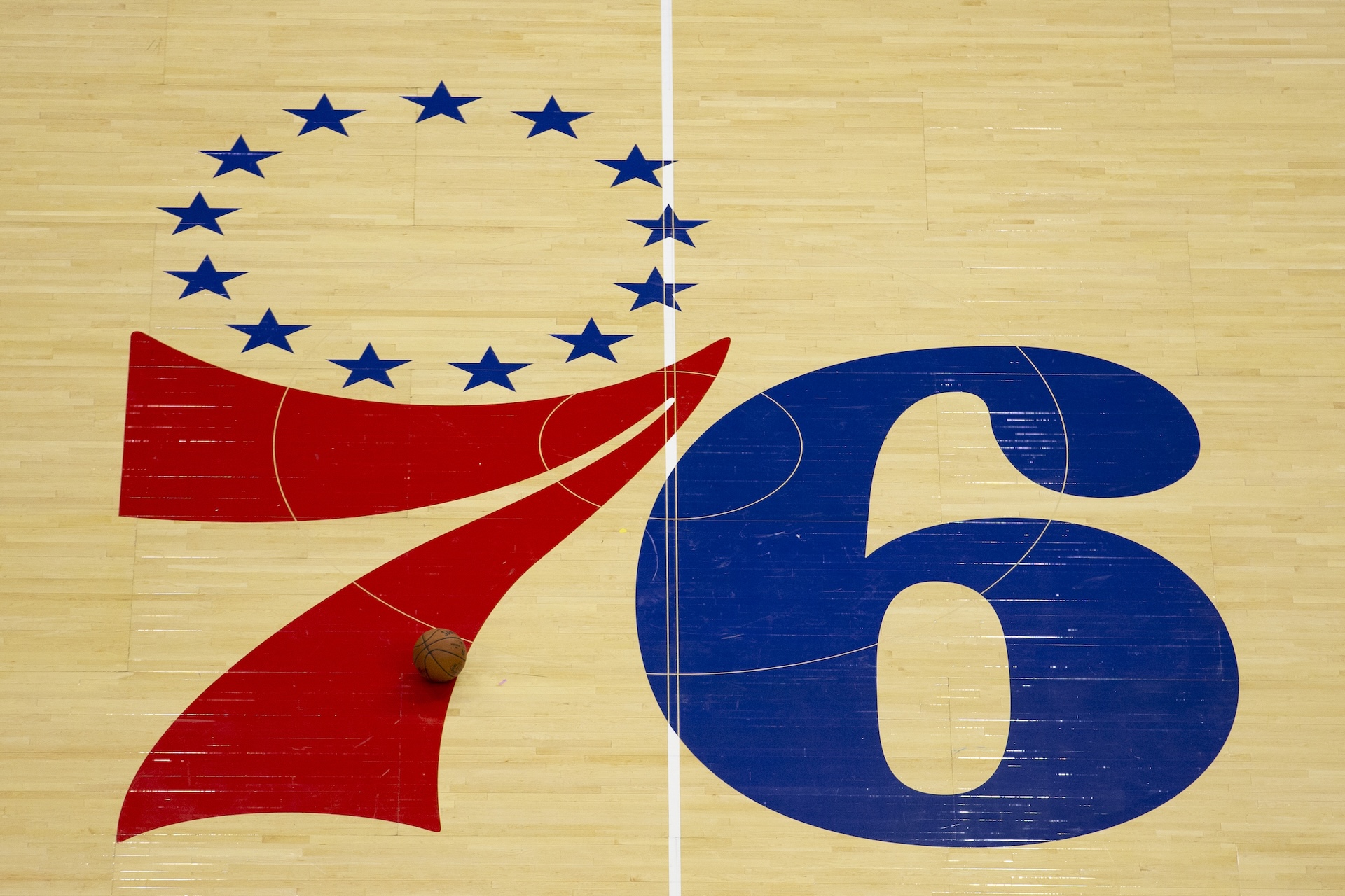 Philadelphia 76ers logo on a basketball court, featuring a red and blue "76" with stars above. A basketball rests on the floor.