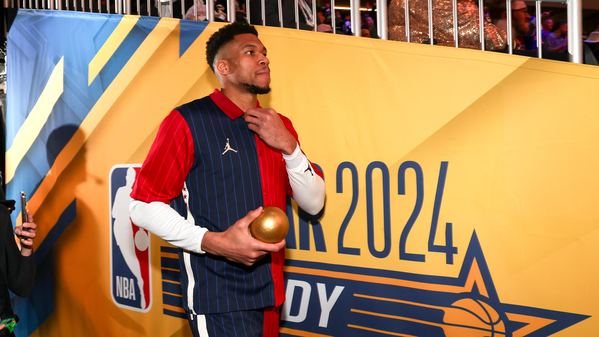 Giannis Antetokounmpo holding a golden basketball, standing near a 2024 NBA All-Star Weekend sign.