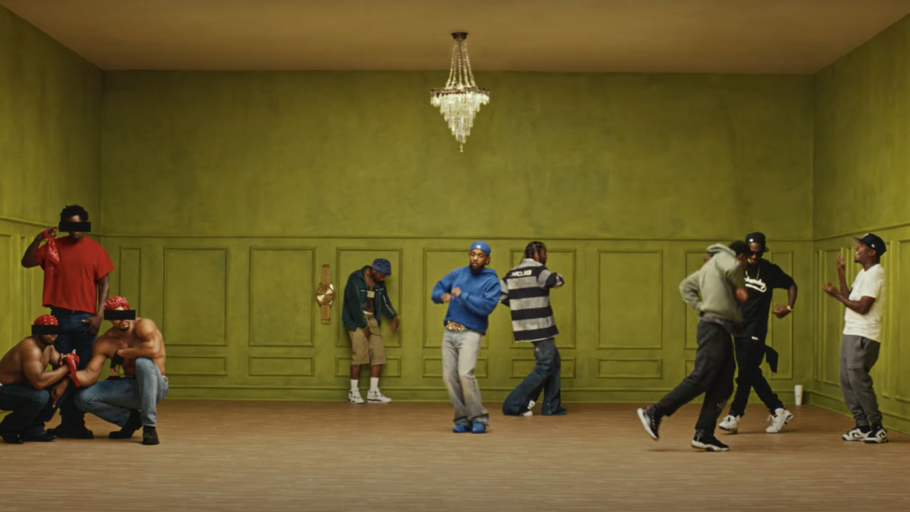 A group of people in a green room, some dancing and others standing or crouching. A chandelier hangs from the ceiling.