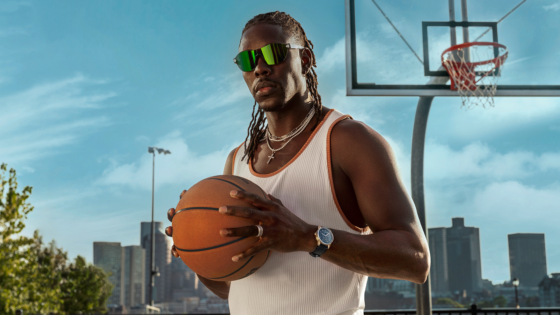 A man wearing sunglasses and a white tank top holds a basketball on an outdoor court, with a city skyline in the background.
