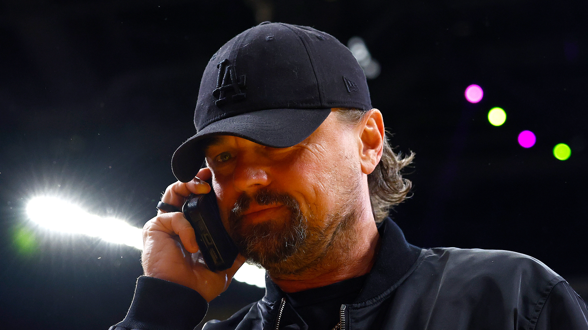 Leonardo DiCaprio wearing a black LA cap and jacket is talking on a phone, with colorful lights in the background.