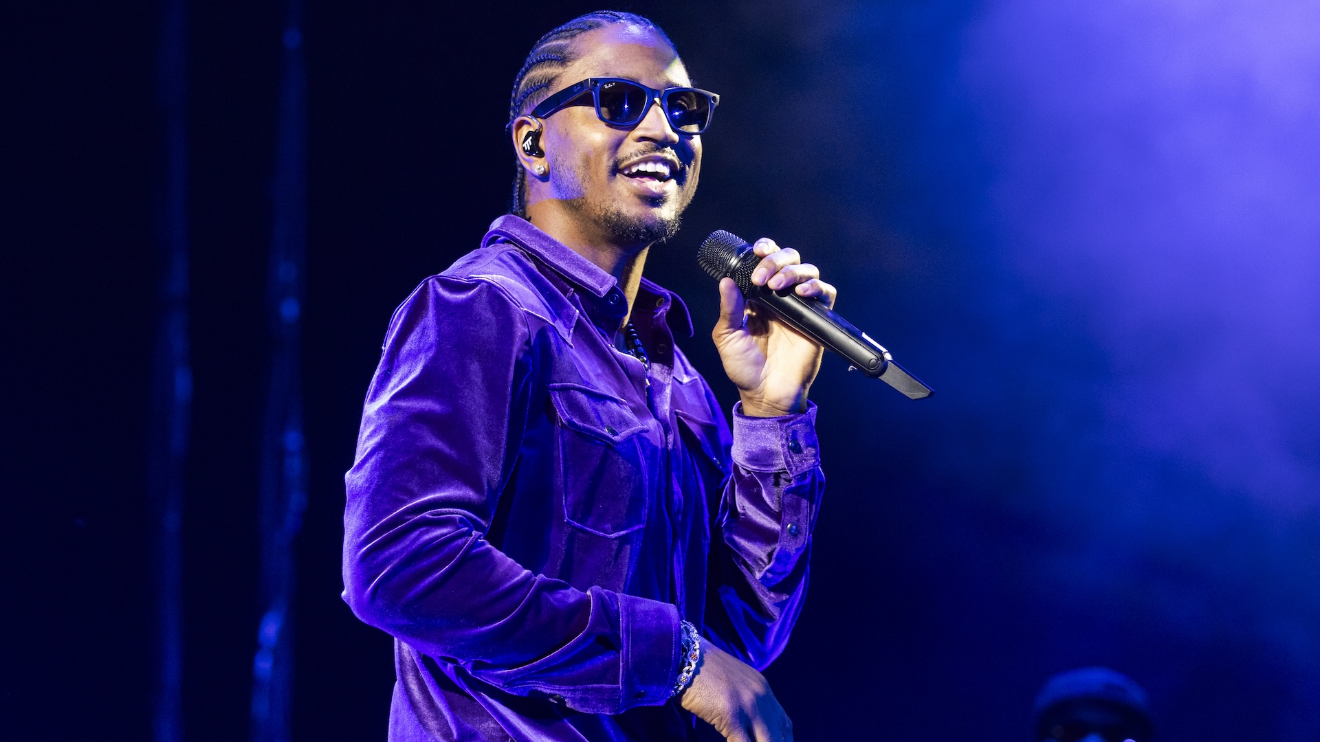 A man wearing sunglasses and a purple velvet shirt performs on stage, holding a microphone, with blue lighting in the background.