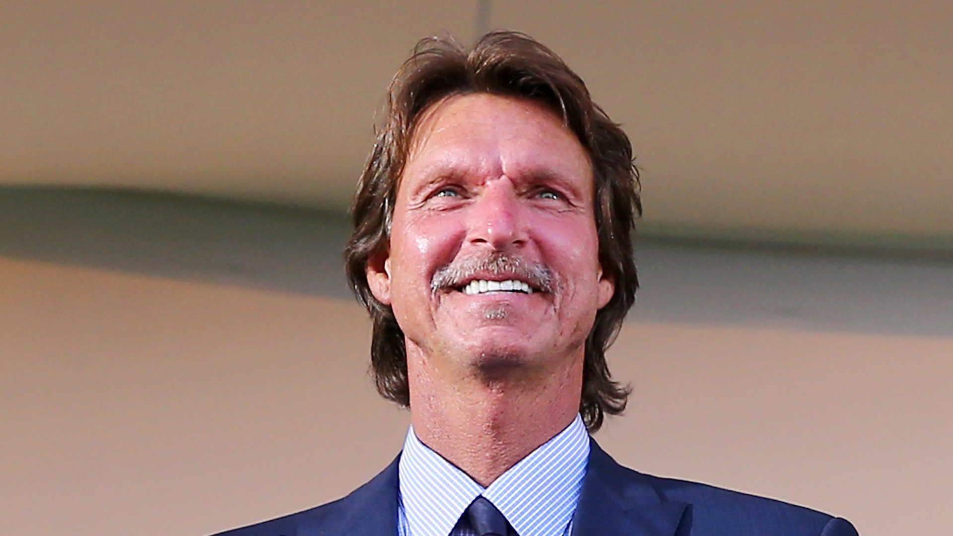 A man with brown hair and a mustache, wearing a suit and tie, smiling.