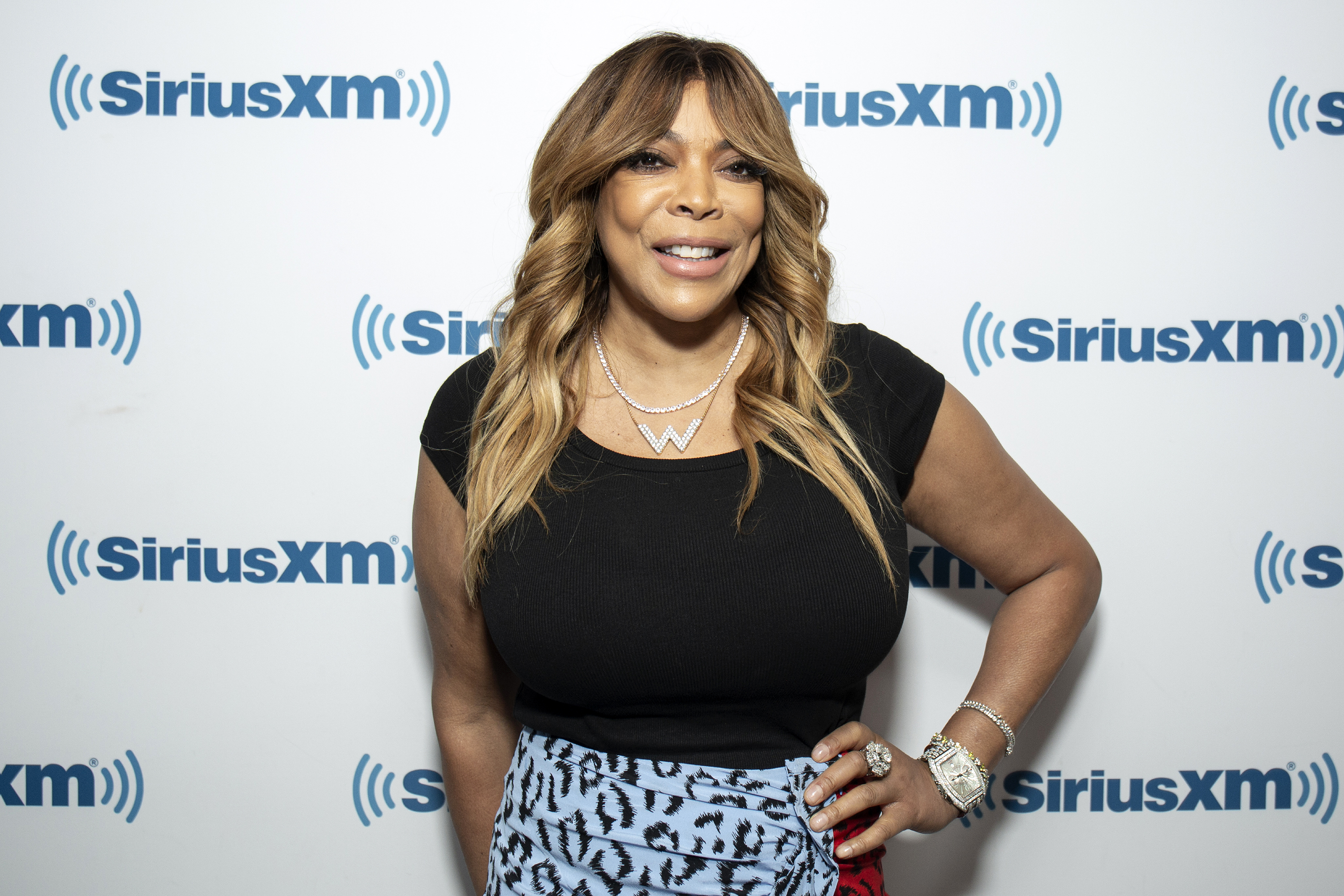 Wendy Williams smiling in front of a backdrop, wearing a black top and patterned skirt, with wavy hair and jewelry.
