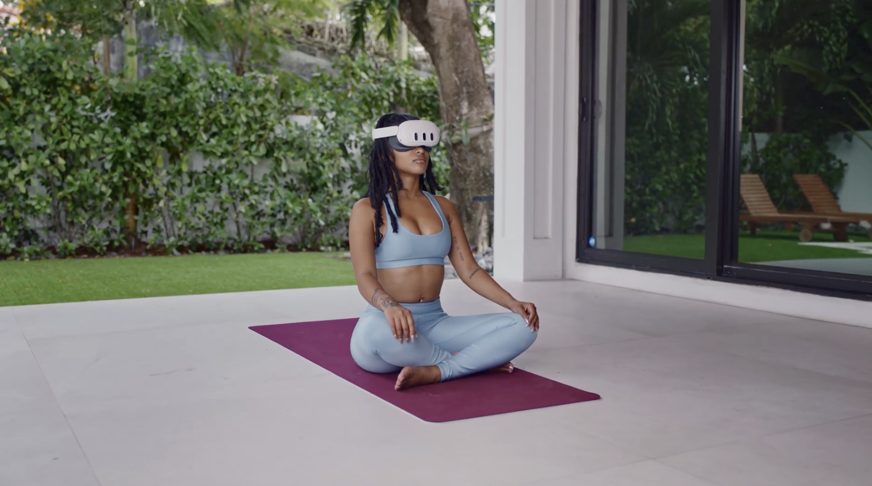 A woman in a blue workout outfit sits cross-legged on a purple yoga mat, wearing a VR headset, in a modern, open space with greenery outside.