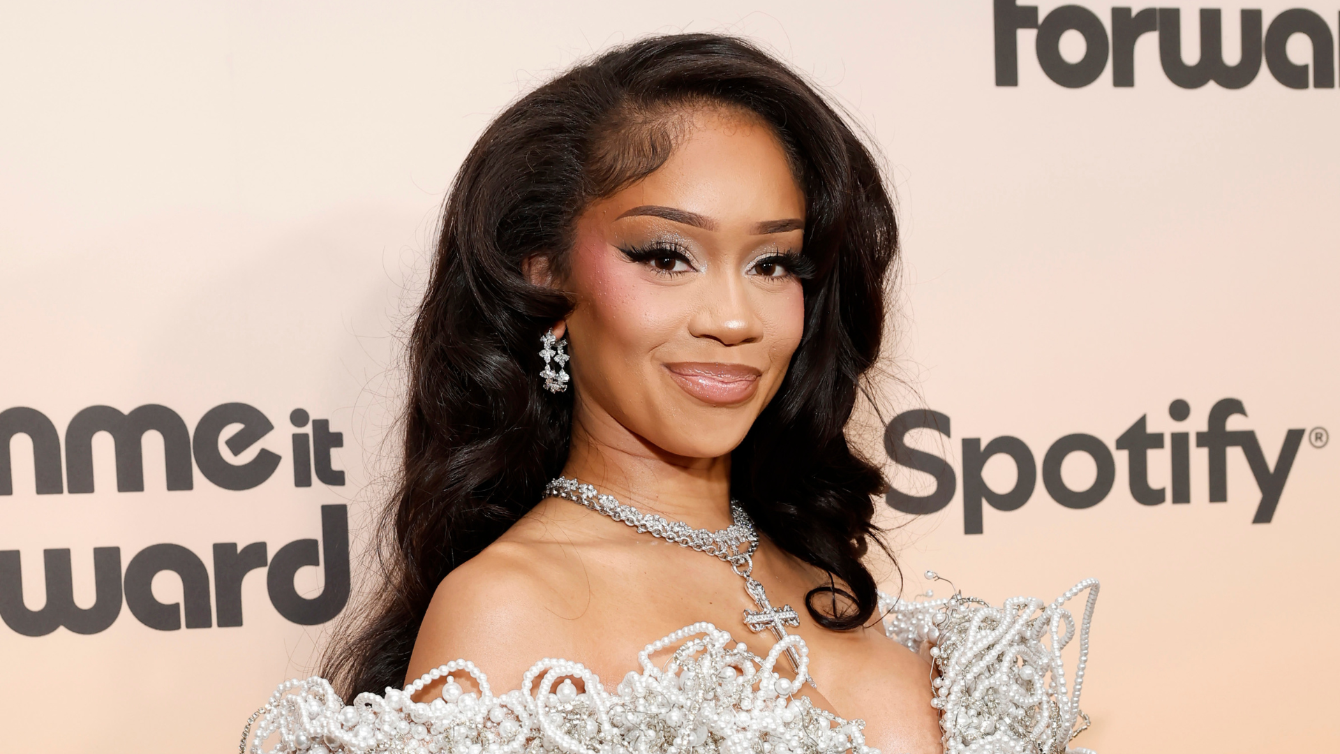 Saweetie at an event, wearing a glamorous silver outfit and jewelry, with long wavy hair and sparkling earrings.