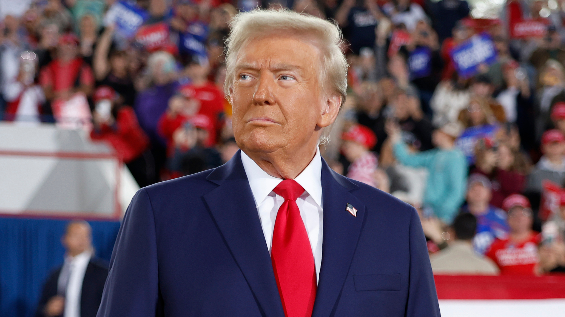 Donald Trump in a navy suit and red tie at a rally, with a crowd in the background holding signs.