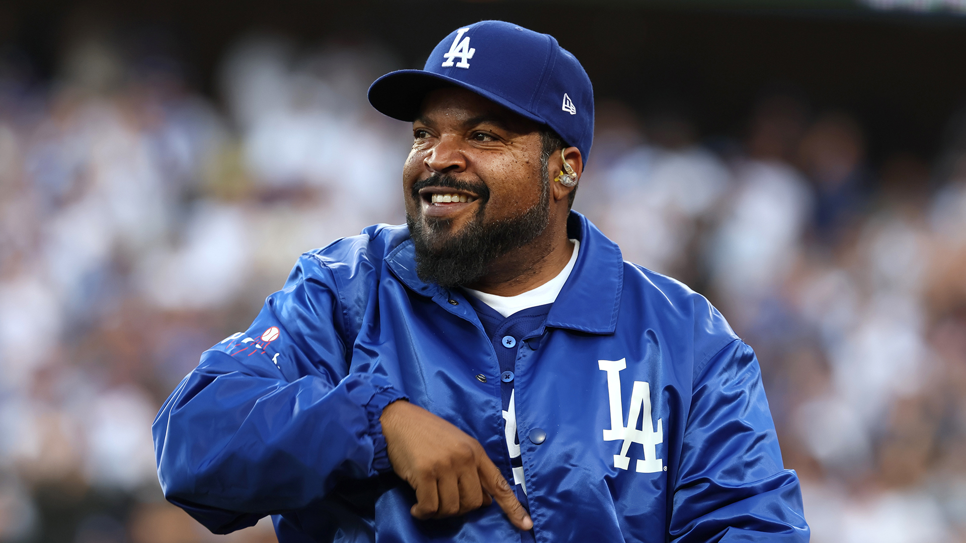 Ice Cube at a Los Angeles Dodgers game, wearing a blue Dodgers jacket and cap, smiling and pointing.