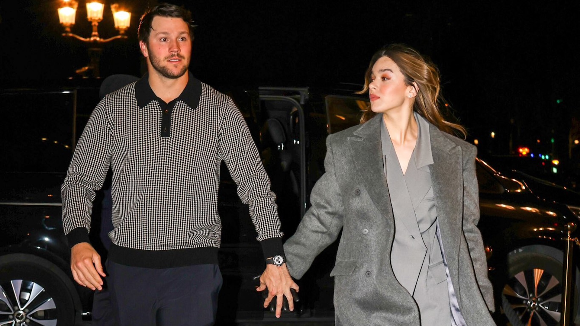 Josh Allen and Hailee Steinfeld walking hand in hand at night. The man wears a patterned sweater, and the woman wears a gray coat.