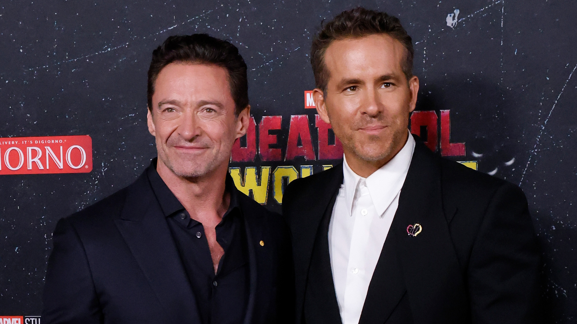 Hugh Jackman and Ryan Reynolds pose together at a "Deadpool" event, both wearing dark suits and smiling.
