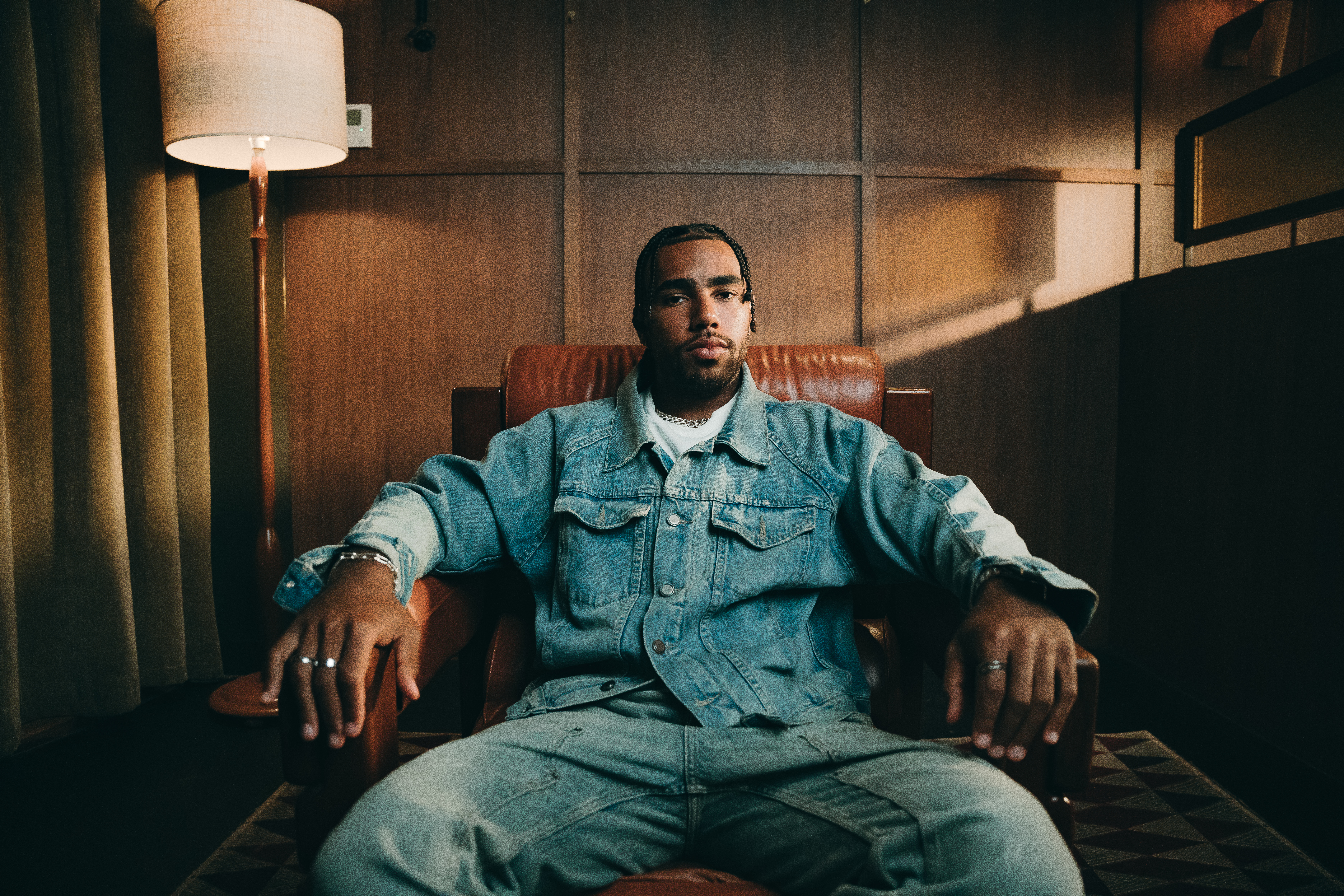 A man sits in a leather chair wearing a denim jacket and jeans, with a relaxed posture. The setting is warmly lit with a floor lamp nearby.