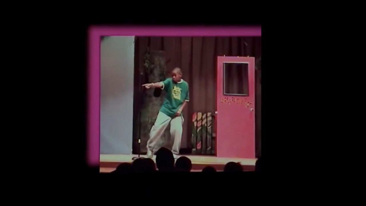 Tyler, The Creator in a green shirt and gray pants performs on stage with a pink backdrop and audience silhouettes in the foreground.