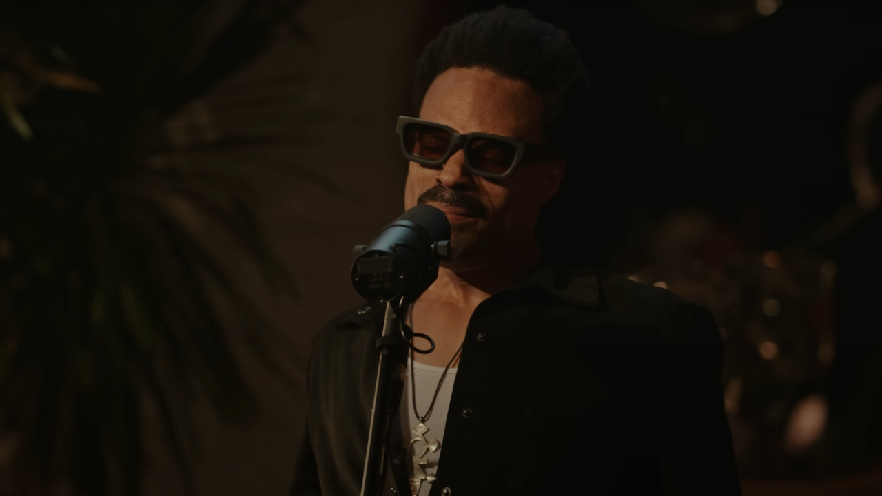 Bilal with sunglasses sings into a microphone in a dimly lit setting.