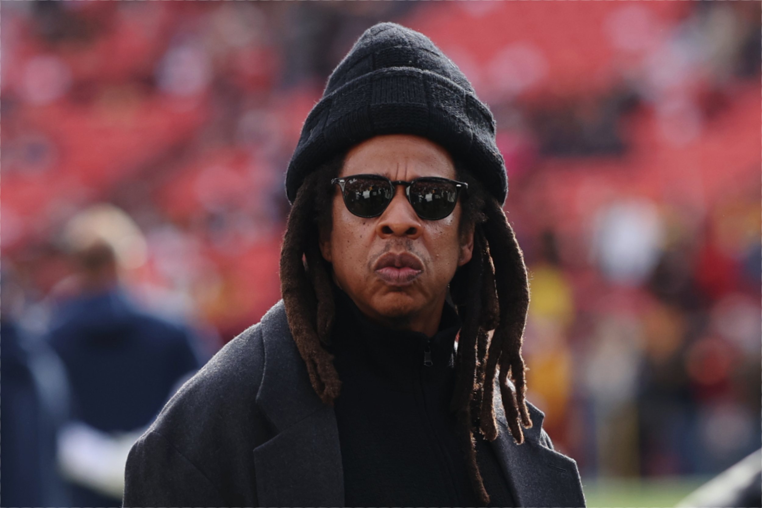 Jay-Z wearing sunglasses, a black beanie, and a dark coat stands outdoors with a blurred crowd in the background.