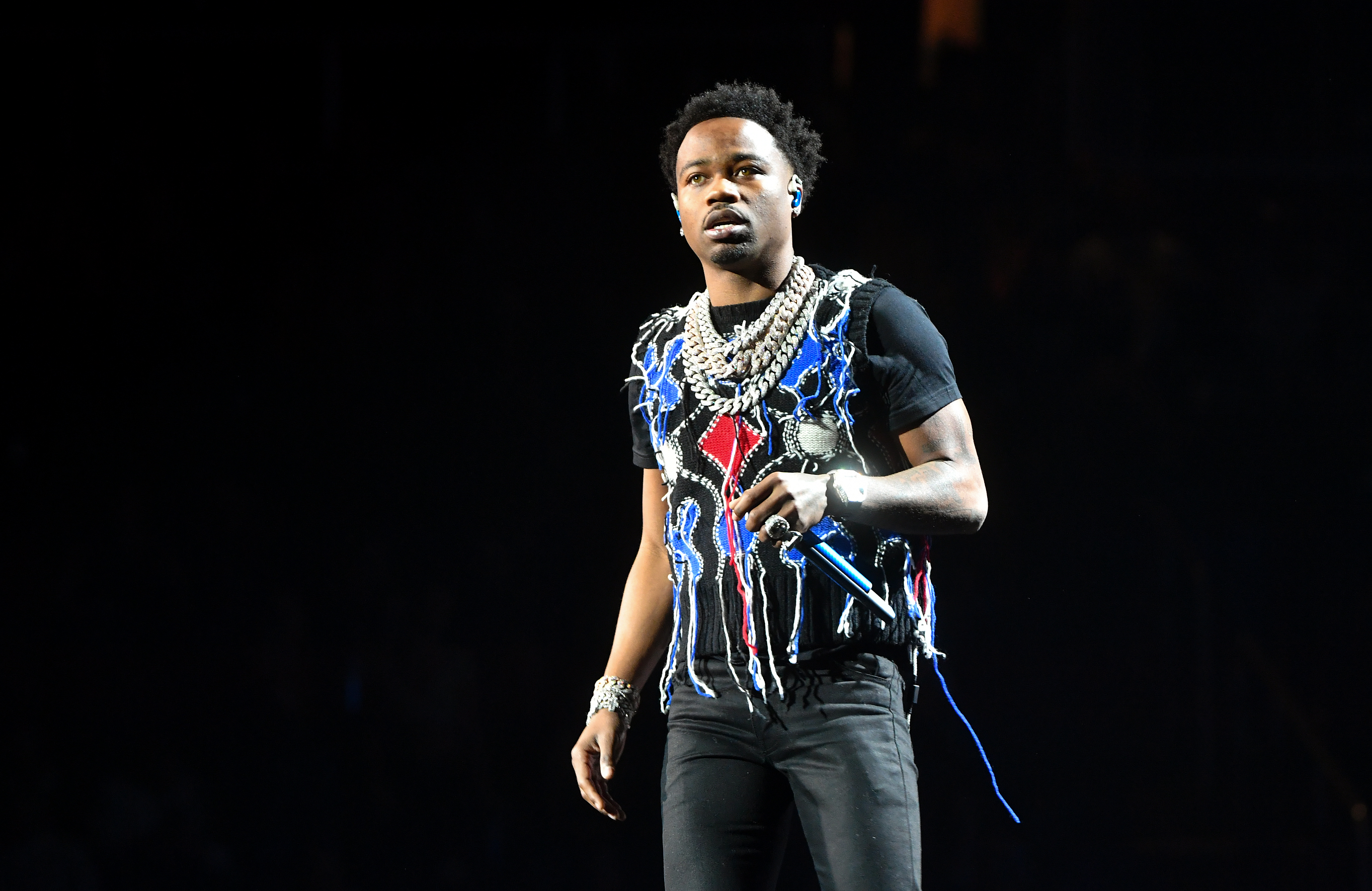 Roddy Ricch performing on stage, wearing a black outfit with colorful patterns and multiple chains, holding a microphone.