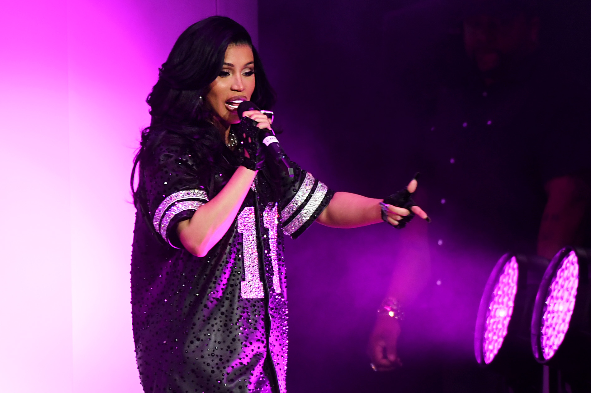 Cardi B performing on stage in a sparkling jersey with the number 11, surrounded by purple lighting.