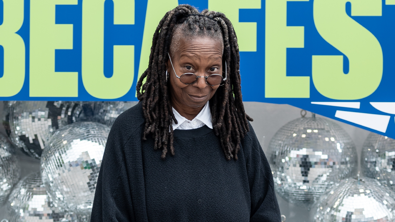 Whoopi Goldberg in front of disco balls and a festival sign, wearing glasses and a black sweater.