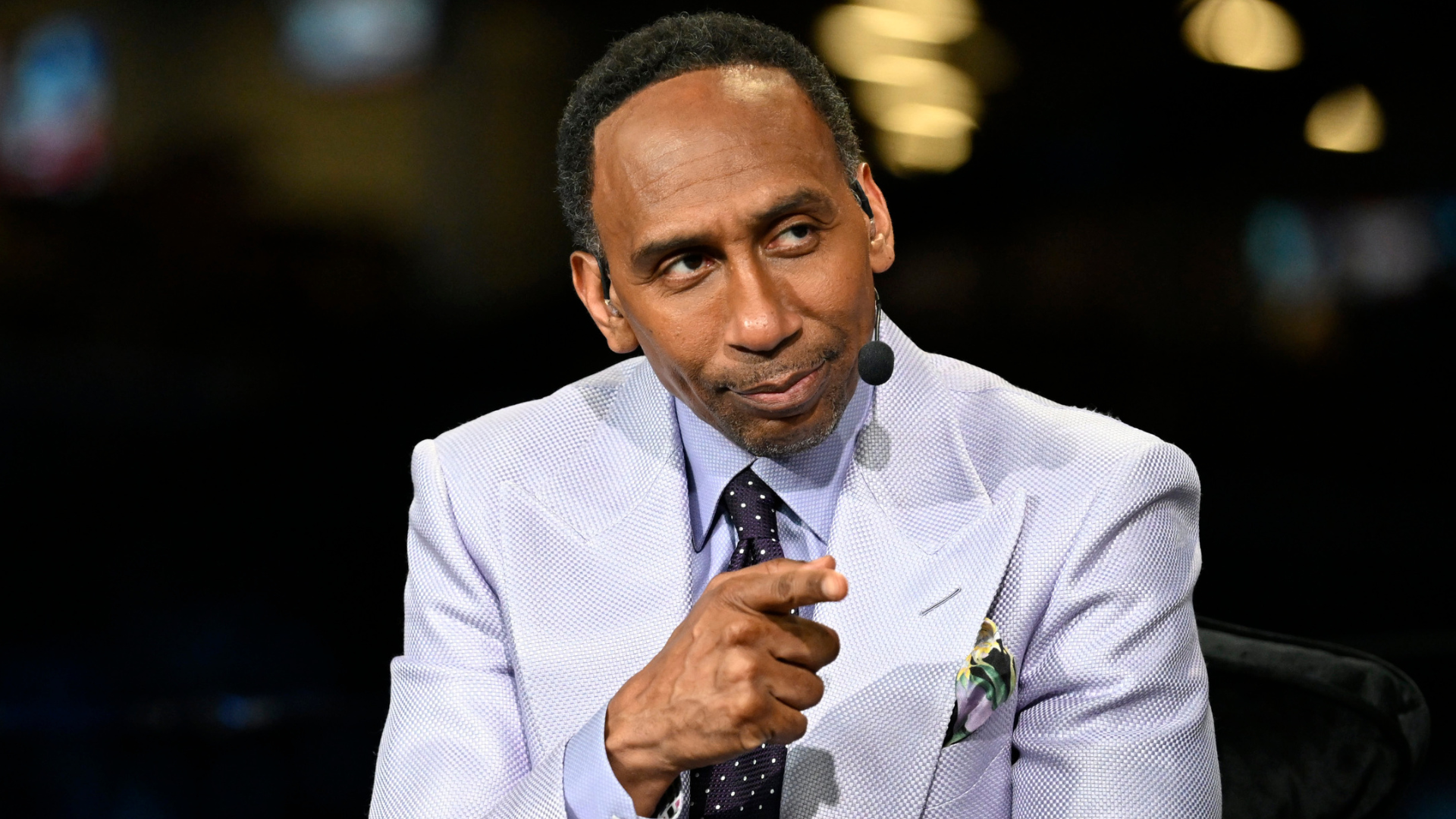 A man in a light purple suit gestures while speaking on a microphone headset, against a blurred background.