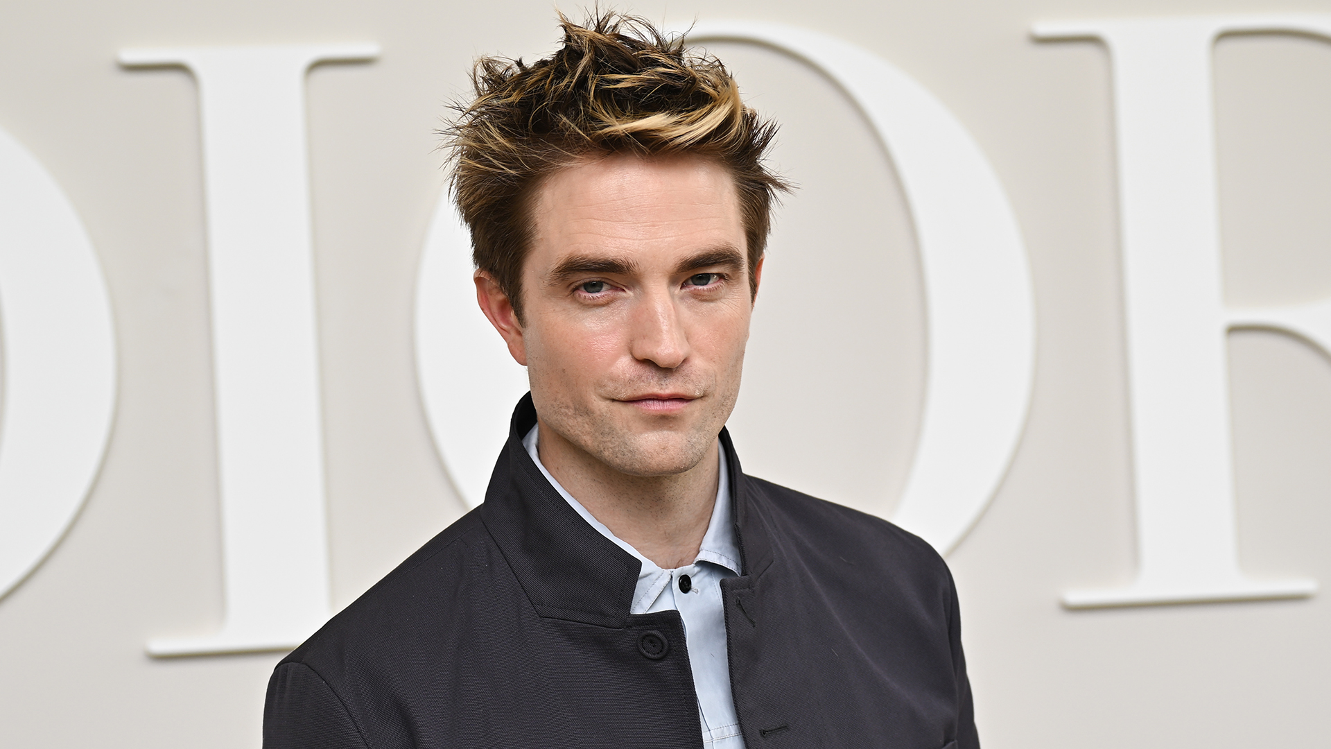 Robert Pattinson at a Dior event, wearing a dark jacket and light shirt, with a neutral expression against a Dior backdrop.