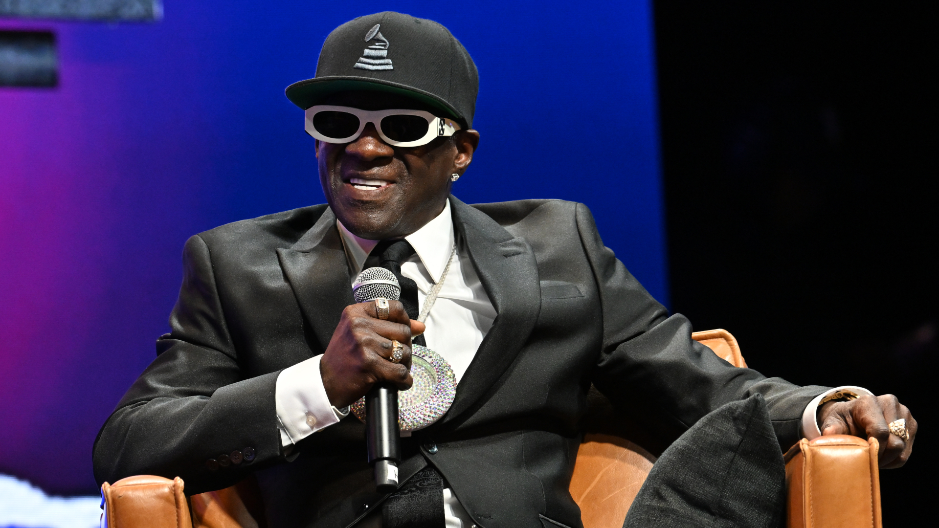 Flavor Flav in a suit, wearing sunglasses and a large clock necklace, holding a microphone, seated on a stage.
