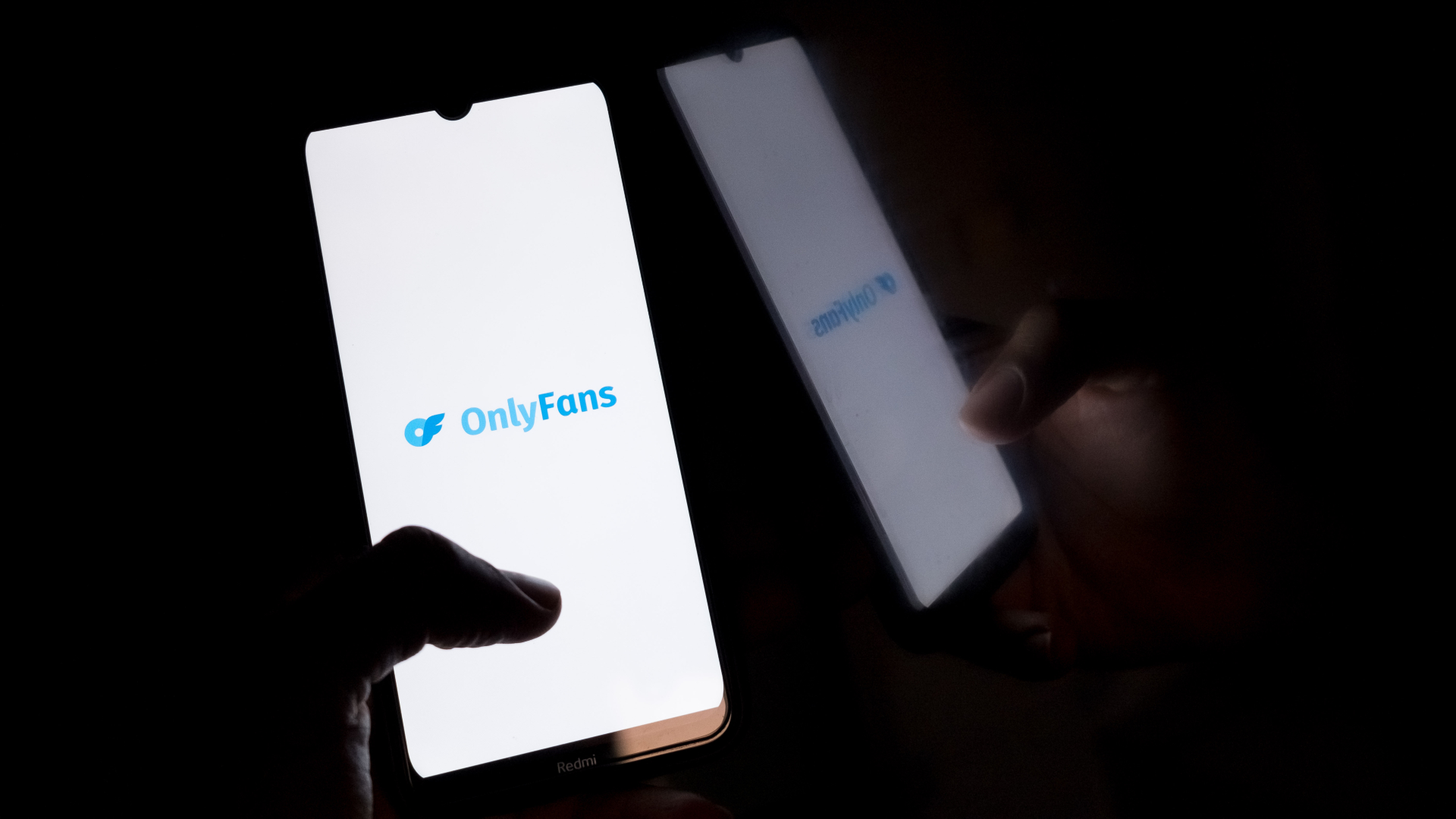 A smartphone displaying the OnlyFans logo on the screen, held by a hand in a dark setting.