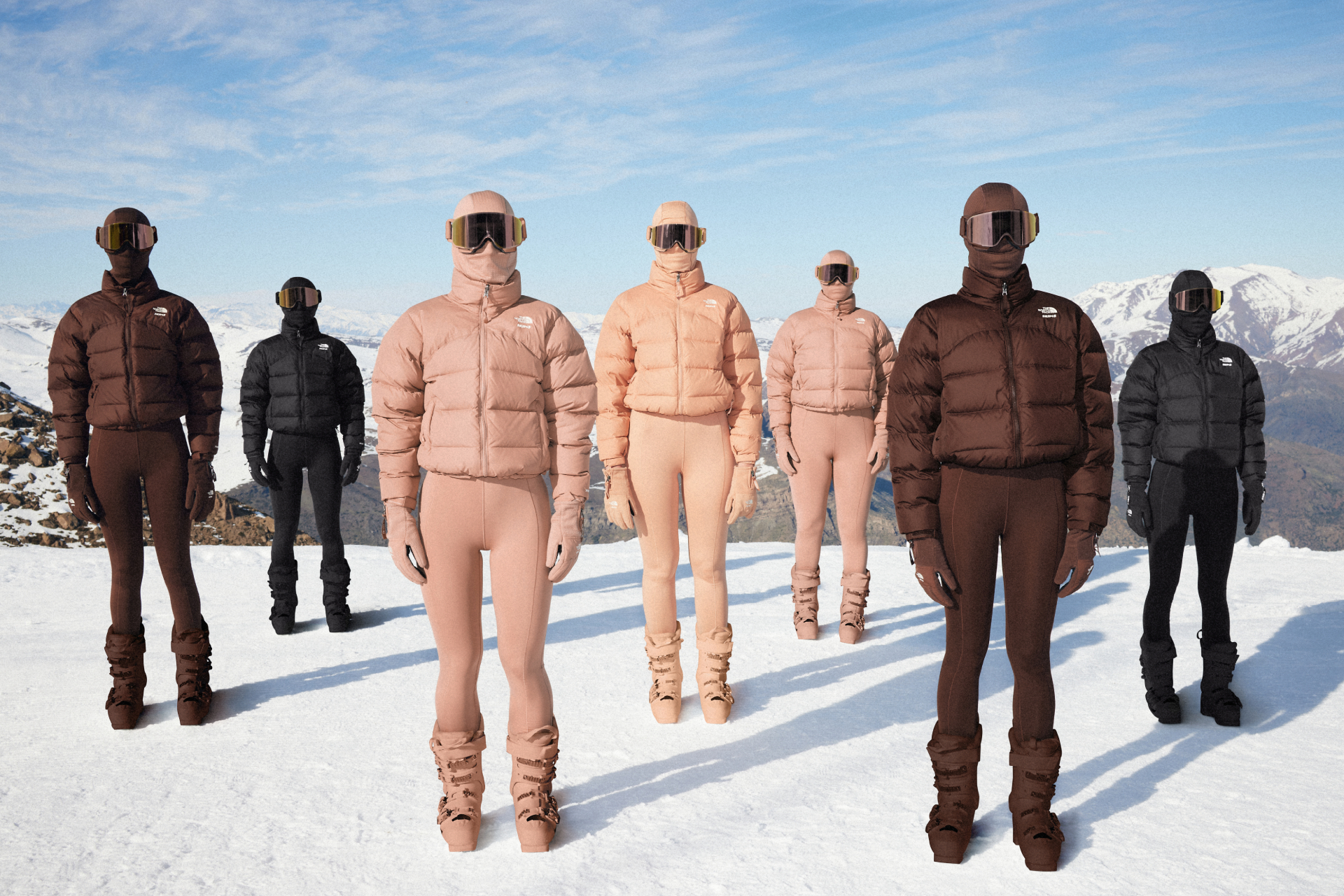 A group of people in matching ski outfits and goggles stand on a snowy mountain, wearing puffy jackets and boots in various earth tones.